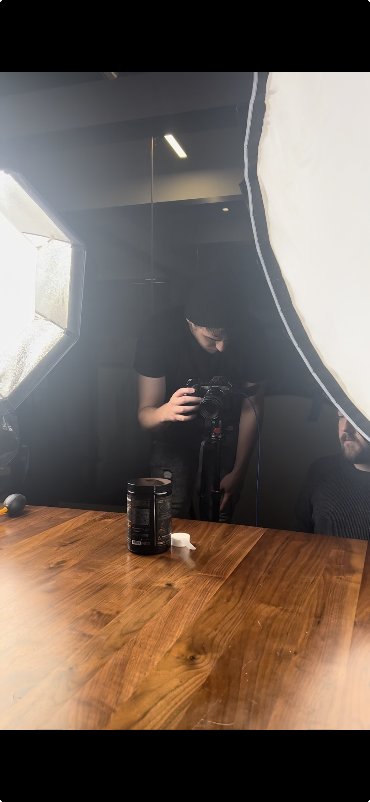 A person taking a photo through a camera mounted on a tripod, in a room with studio lighting and a wooden table, another person partially visible on the right.