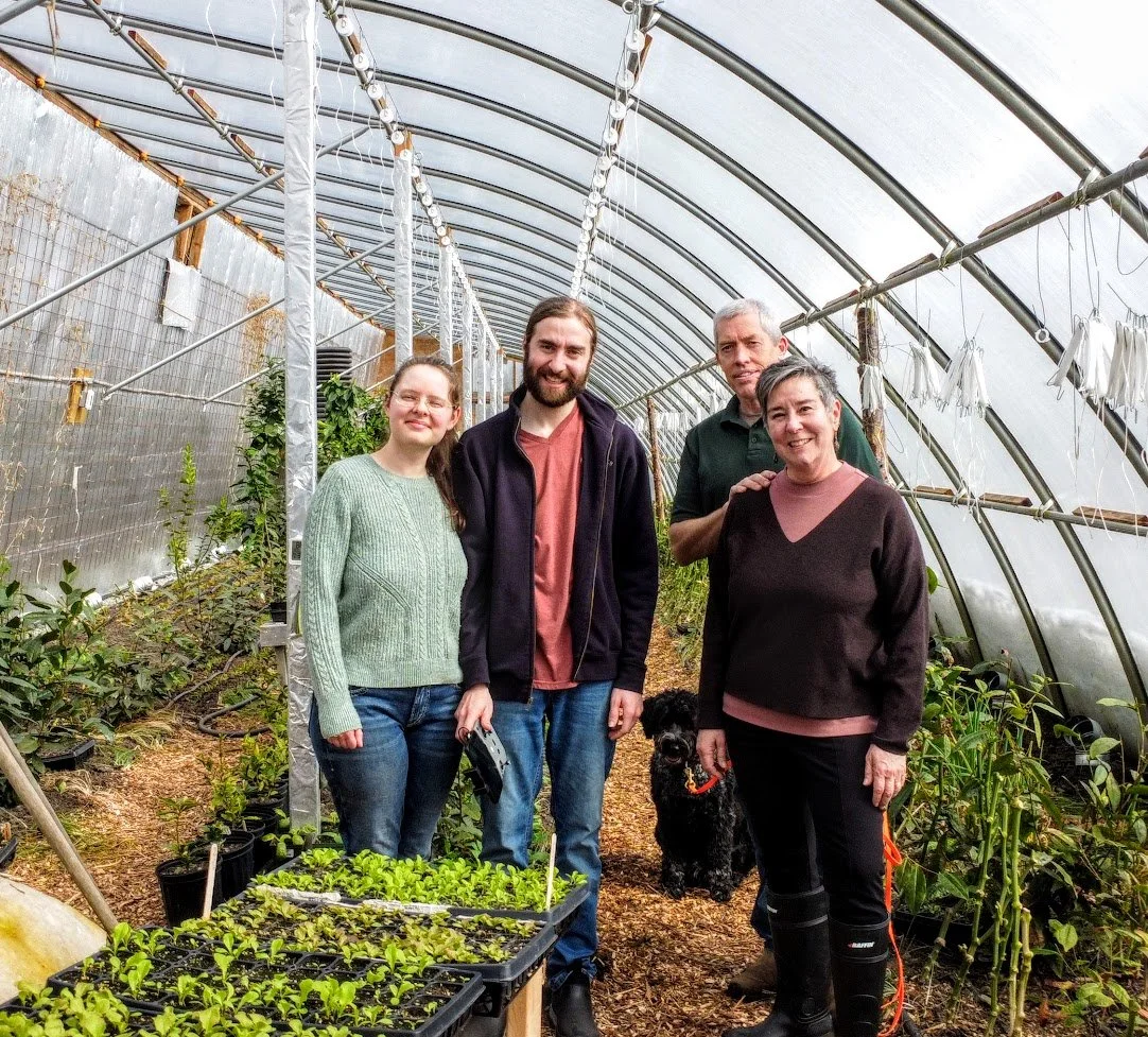 The two founders of Coastal Grove Farm and their family, a group of four passionate sustainable organic farmers pose for a photo in their greenhouse.