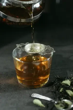 Hot tea being poured into a glass cup on a dark surface, with loose tea leaves and a spoon nearby.