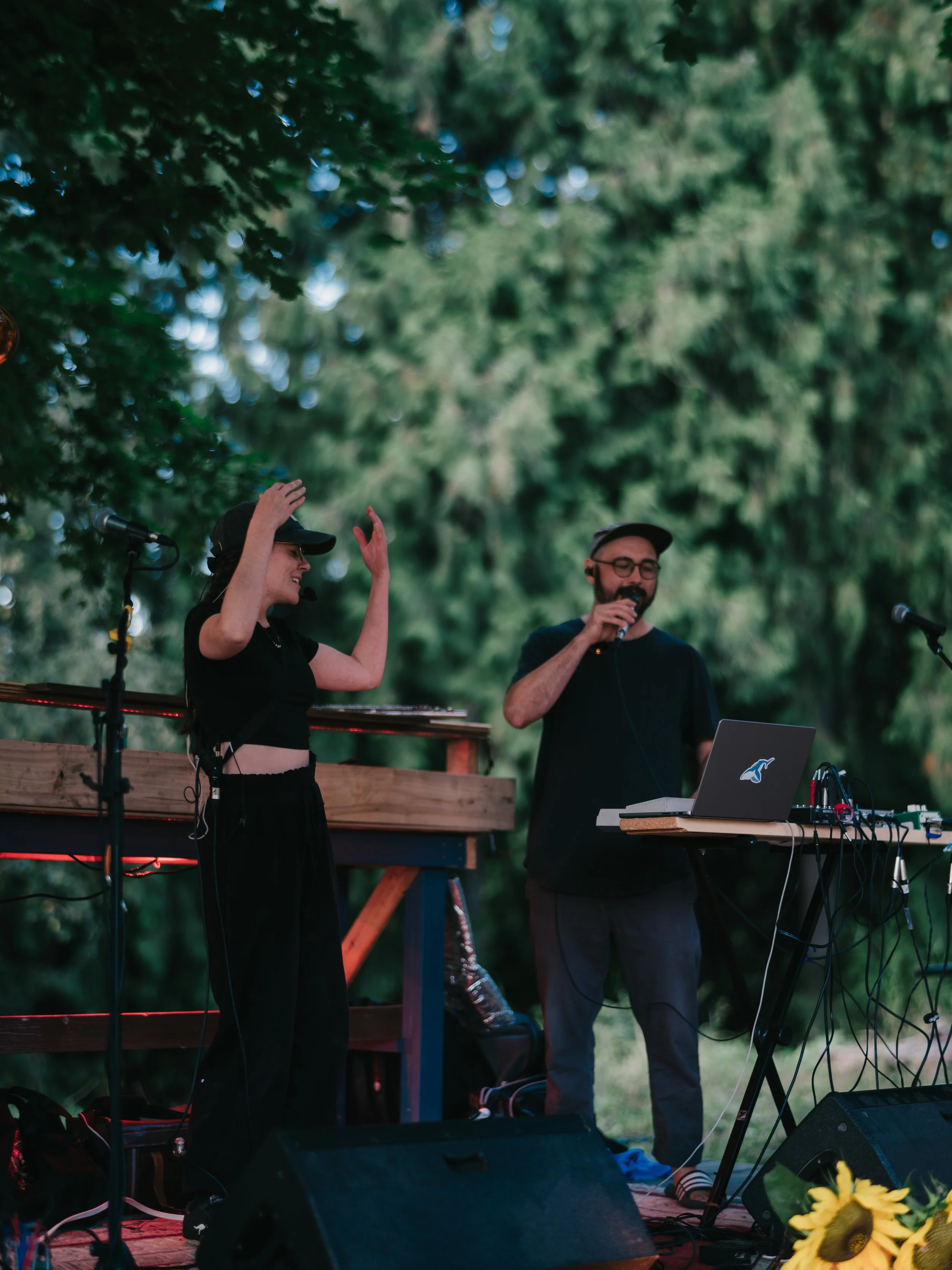 2 people are on stage, there is a woman on the left wearing black pants, a black croptop and grey hat and she is signing. The man is wearing all black and is holding a microphone and singing
