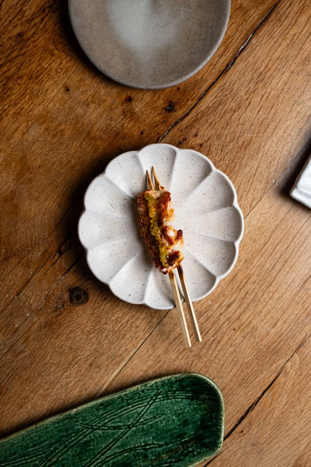 A white scalloped plate with a sliced piece of skewered meat topped with green sauce on a wooden table.