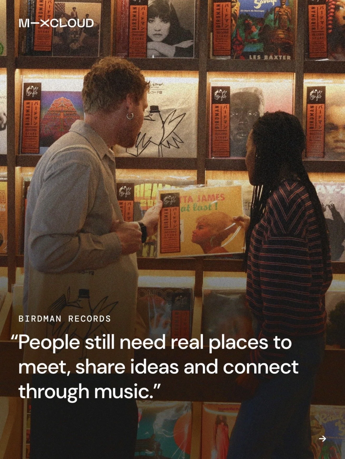 Two people are standing in front of shelves filled with vinyl records at Birdman Records store. One person is holding a record, and the other is looking at their phone. They are engaged in conversation. The background shows colorful album covers and a quote about the importance of real places for connection through music.