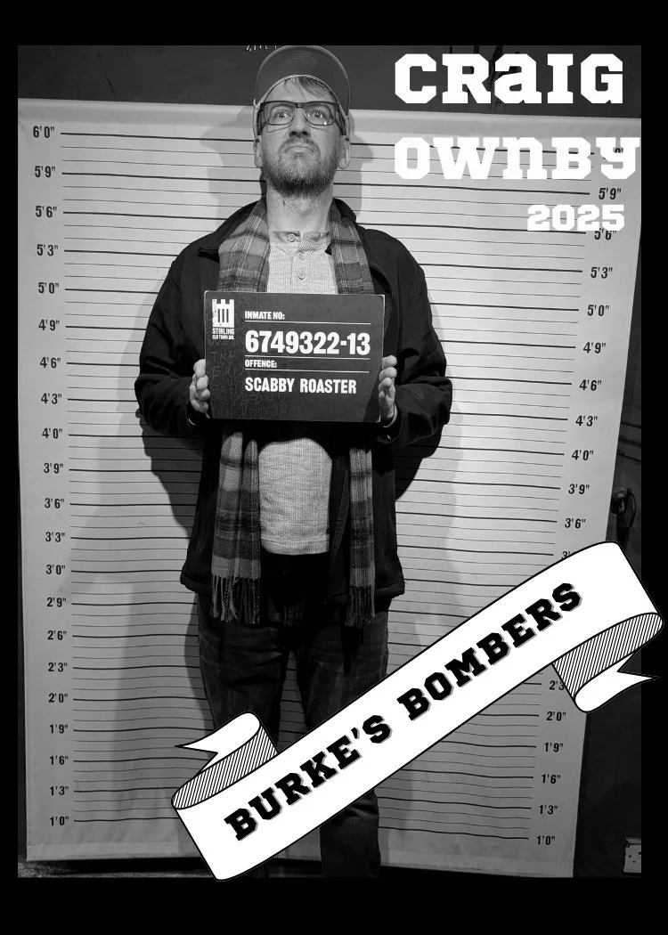 Man with glasses and beard standing in front of a height chart, holding a sign with inmate number 6749322-13, labeled 'Scabby Roaster', at Burke's Bombers event, in black and white.