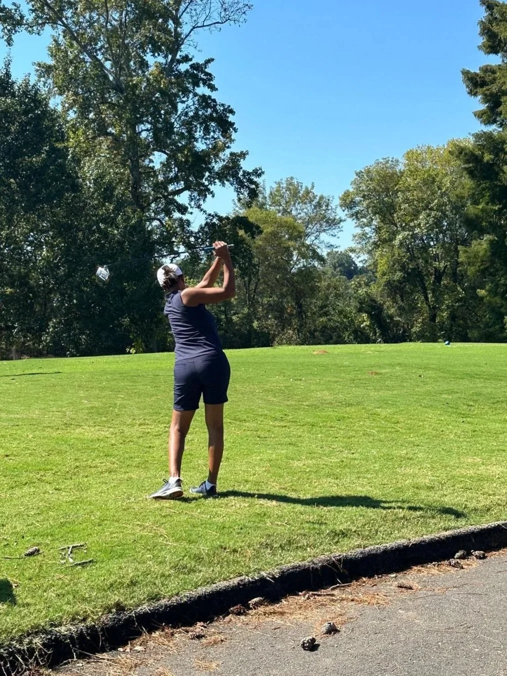 A woman playing golf on a lush green golf course under a clear blue sky, with trees in the background.