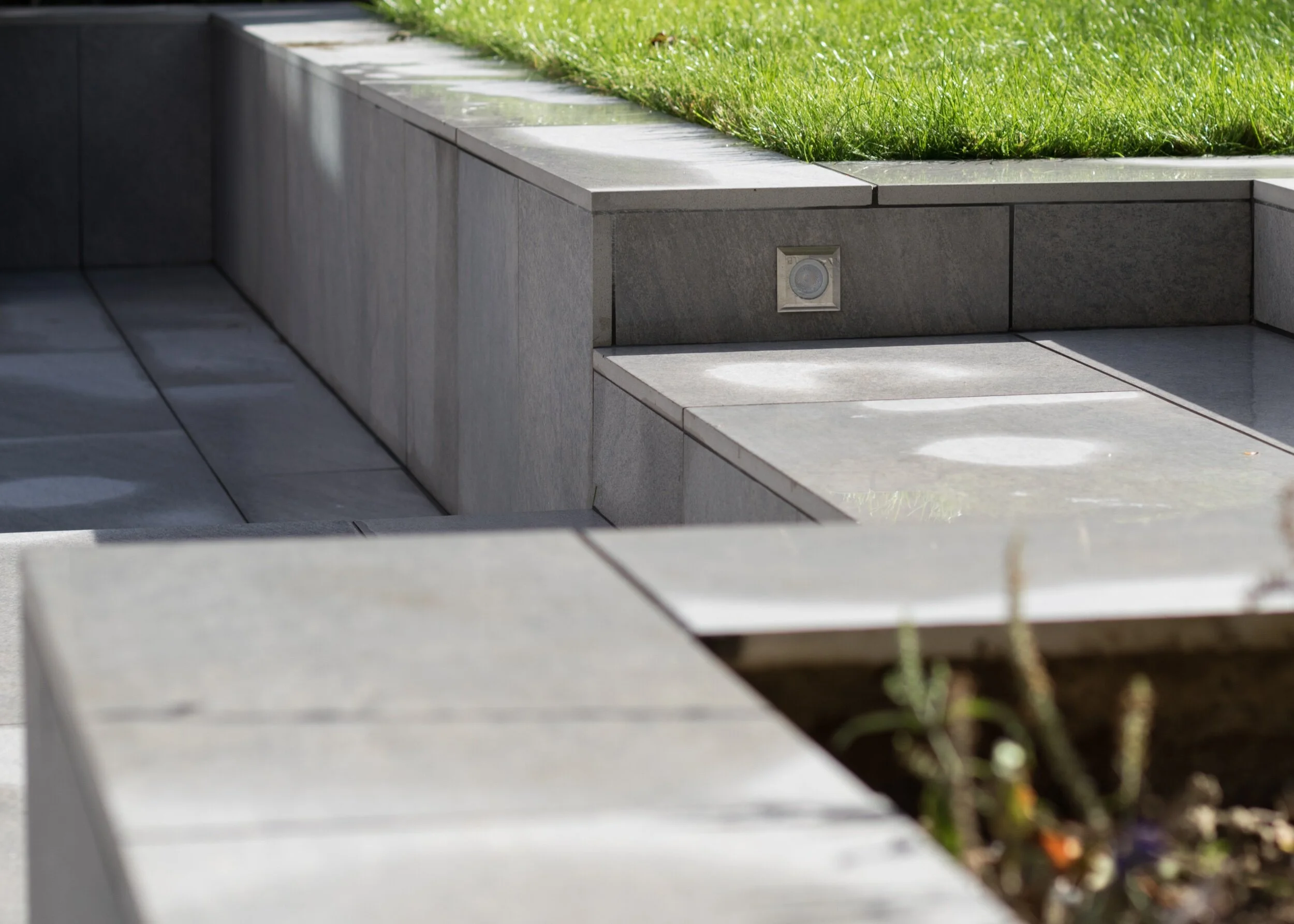 Close-up of modern gray stone outdoor seating area with built-in lighting, surrounding green grass, and a small garden with plants in the foreground.