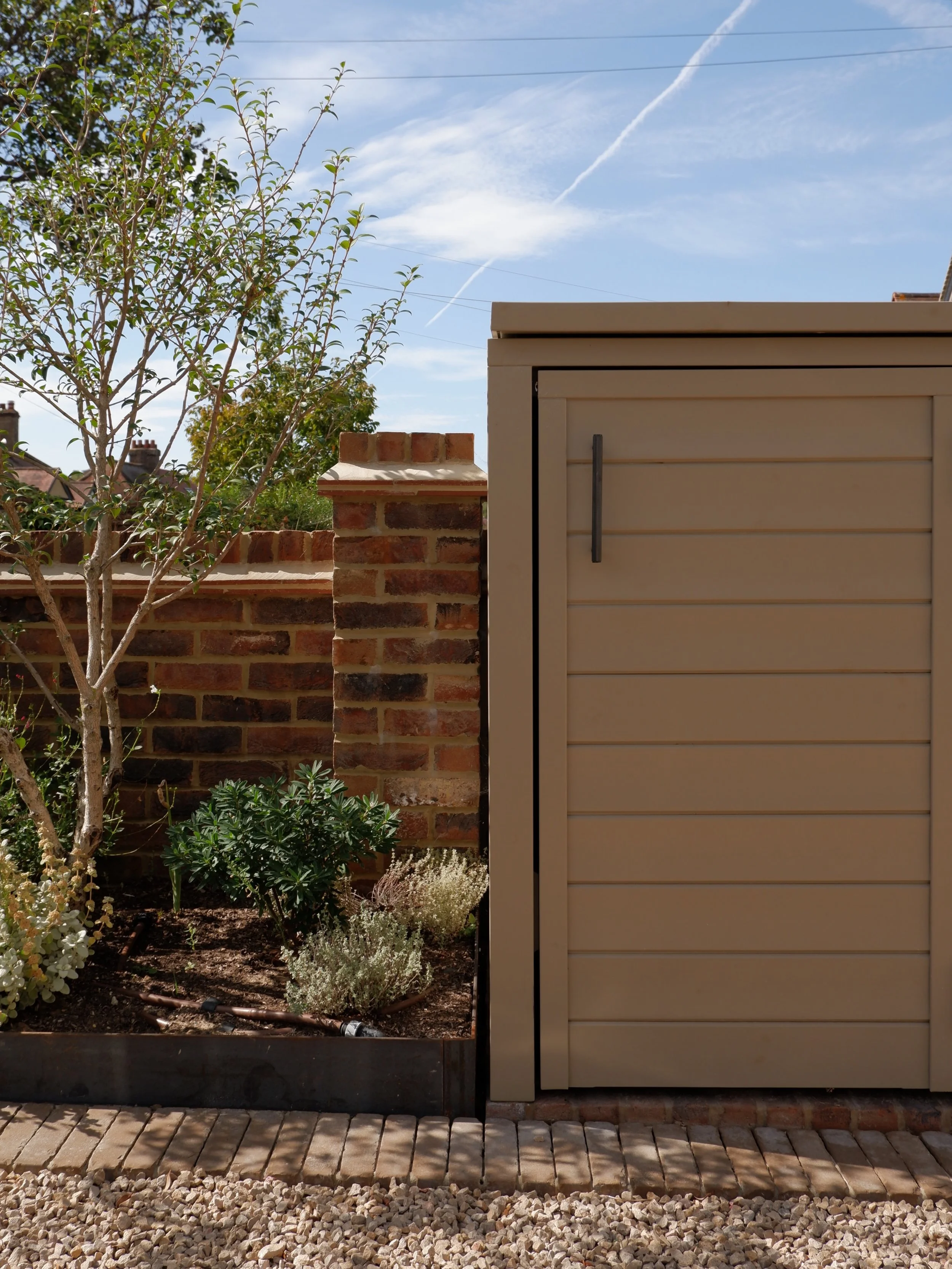 A garden with various green plants and trees, a brick wall, and an outdoor shed or cabinet on the right. The sky is partly cloudy with some contrails visible. Harrington brothers Landscapes Surrey.