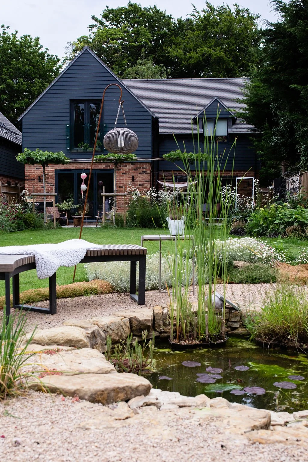 A backyard garden with a pond, plants, and a modern two-story house with dark blue siding and brick accents.