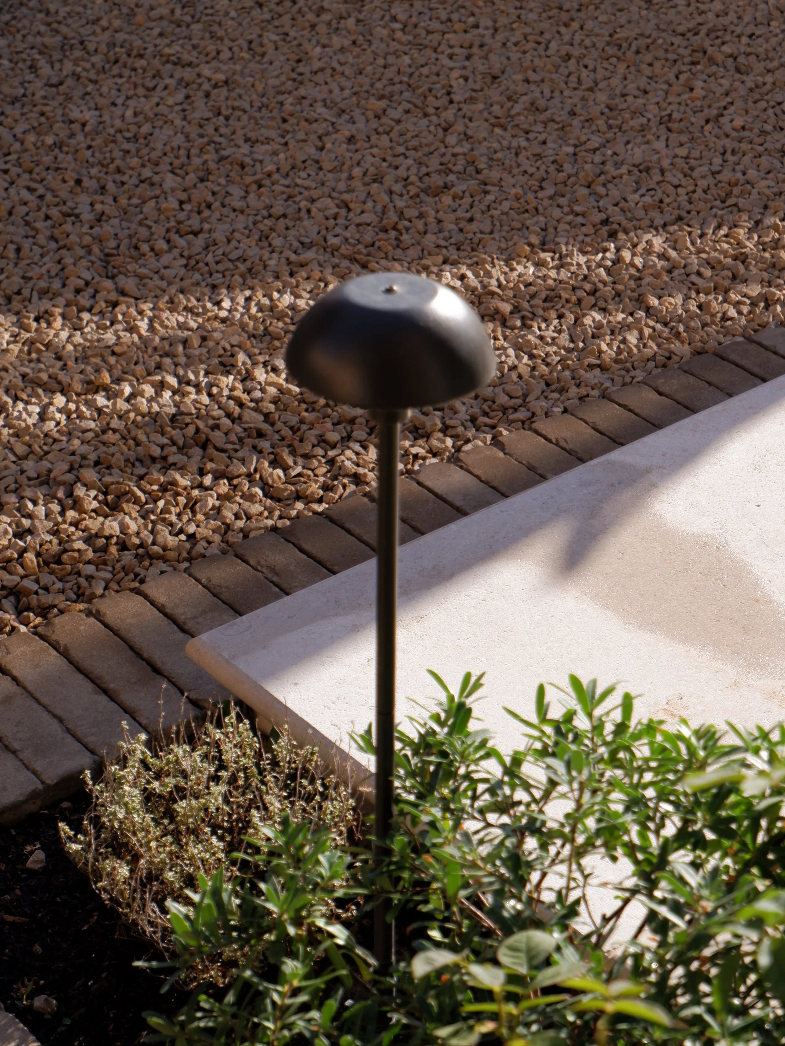 Close-up of a garden bed with a solar-powered garden light, small green plants, mulch, brick border, and concrete walkway. Harrington brothers Landscapes Surrey.