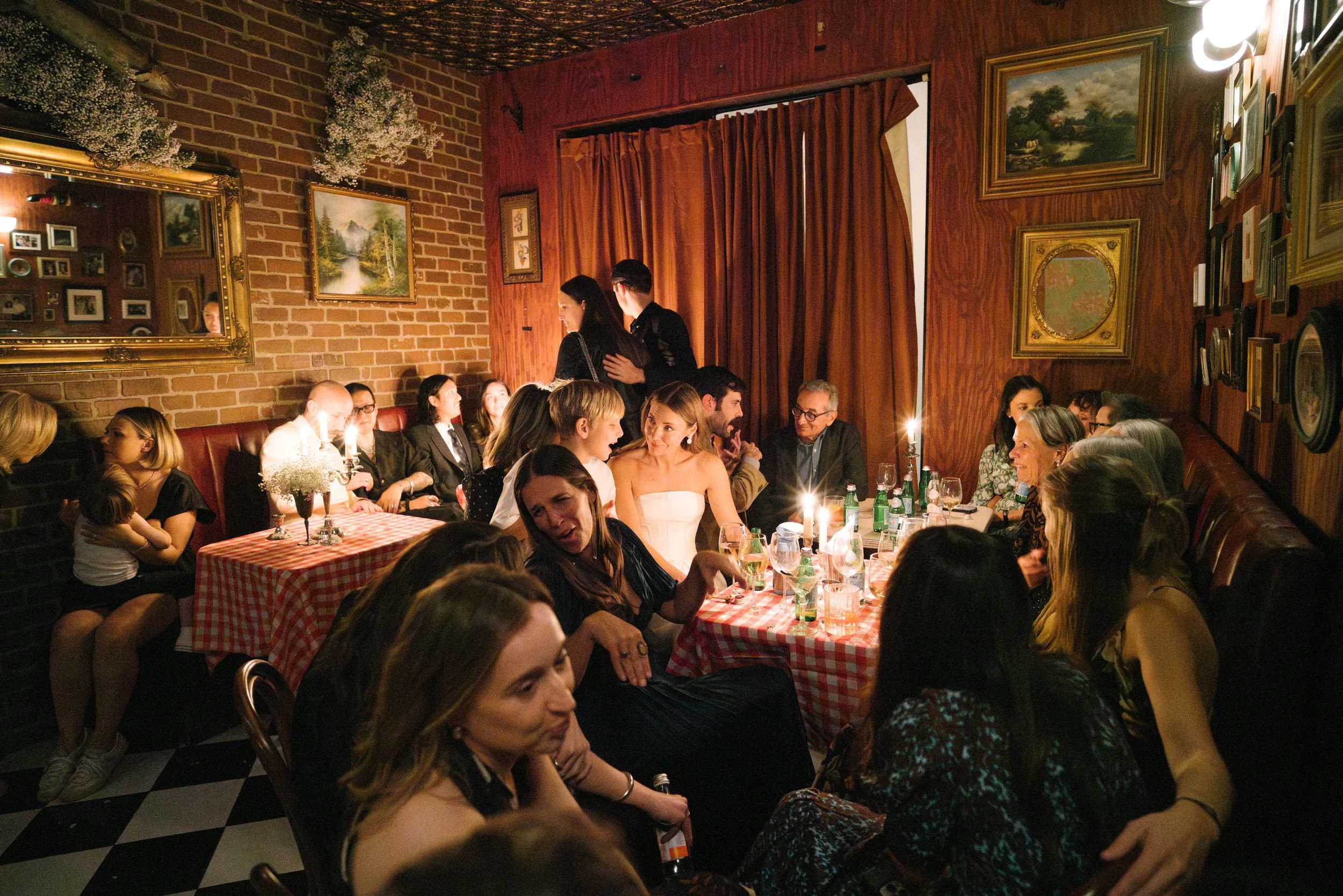 A lively gathering of people seated at a long table in a cozy restaurant or cafe, decorated with vintage paintings and a checkered tablecloth, with warm lighting and a brick wall backdrop.