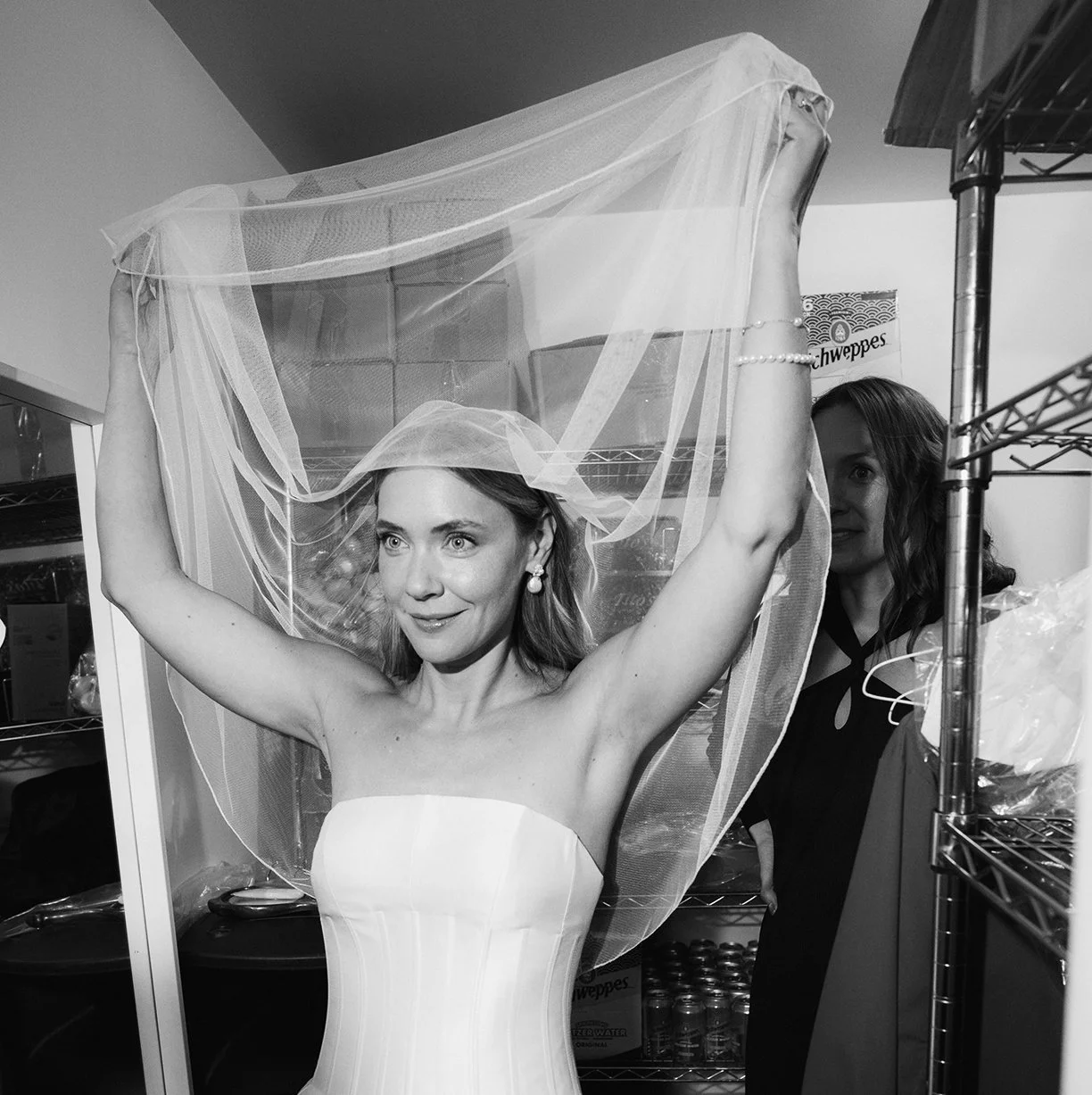 A woman in a wedding dress adjusting her veil in a room with shelves, with another woman partially visible behind her.