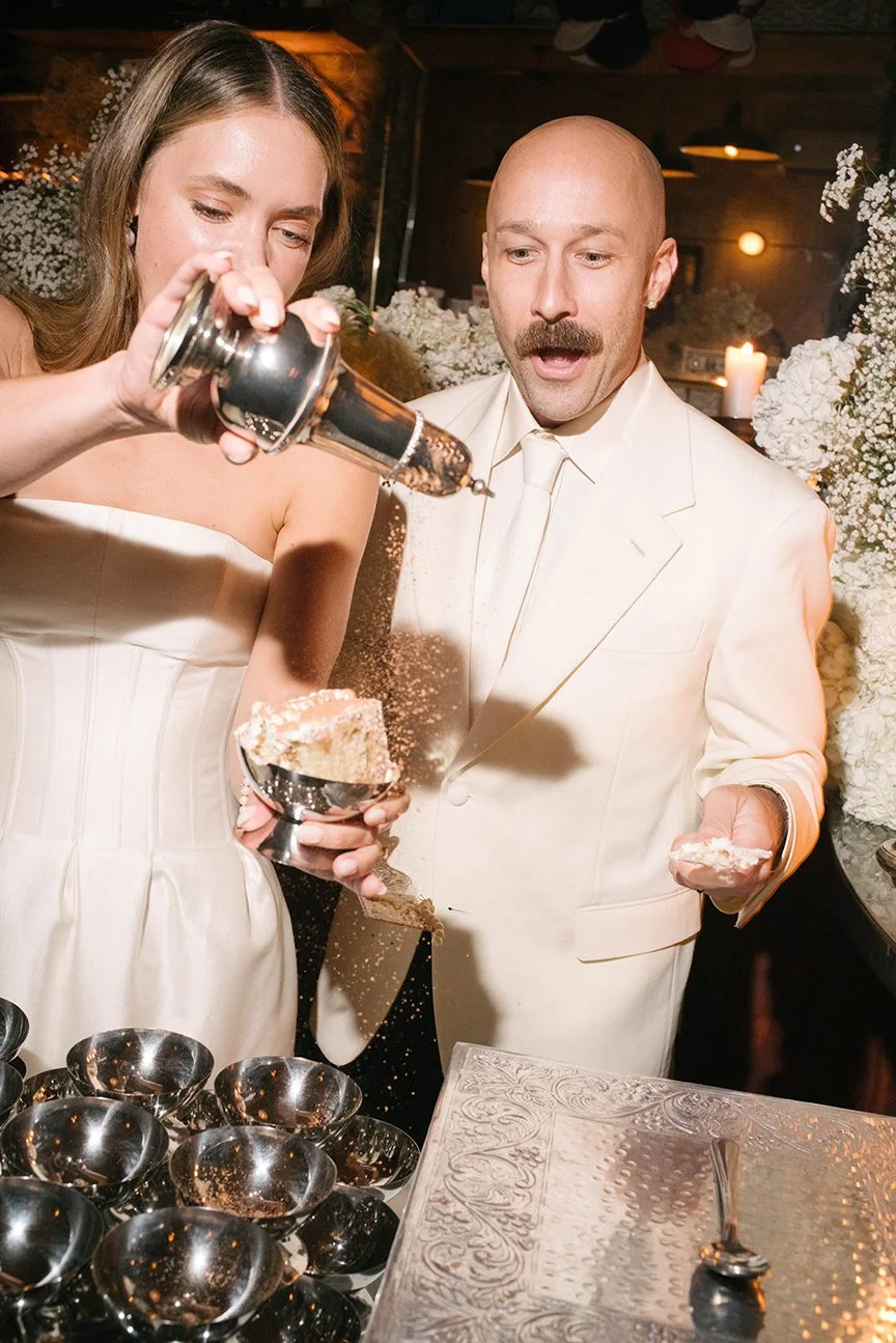 A bride and groom at their wedding celebration, with the bride pouring chocolate or another topping over a dessert while the groom watches surprised, surrounded by candles and flowers.