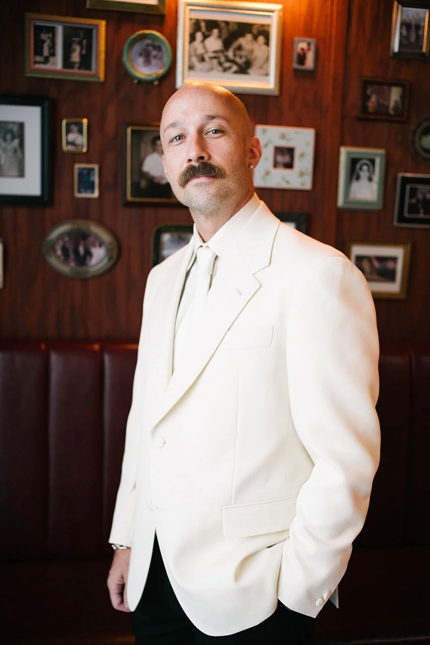 A man with a mustache and a shaved head wearing a white suit jacket and tie, standing in front of a wall decorated with framed photos.