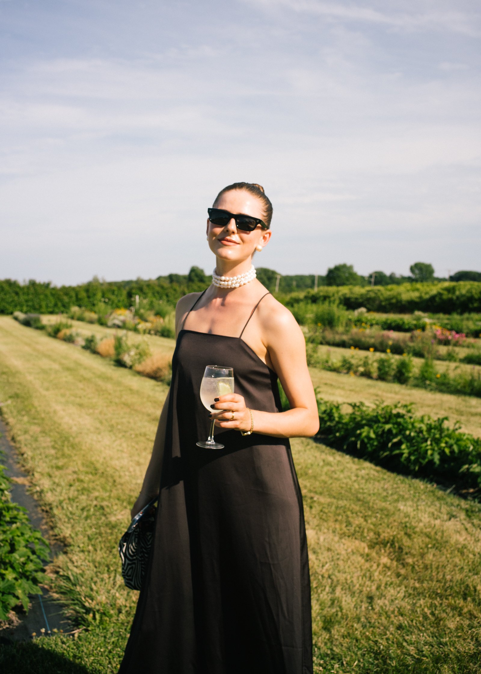 Woman in a black dress holding a glass of wine or cocktail outdoors in a green field under a blue sky.
