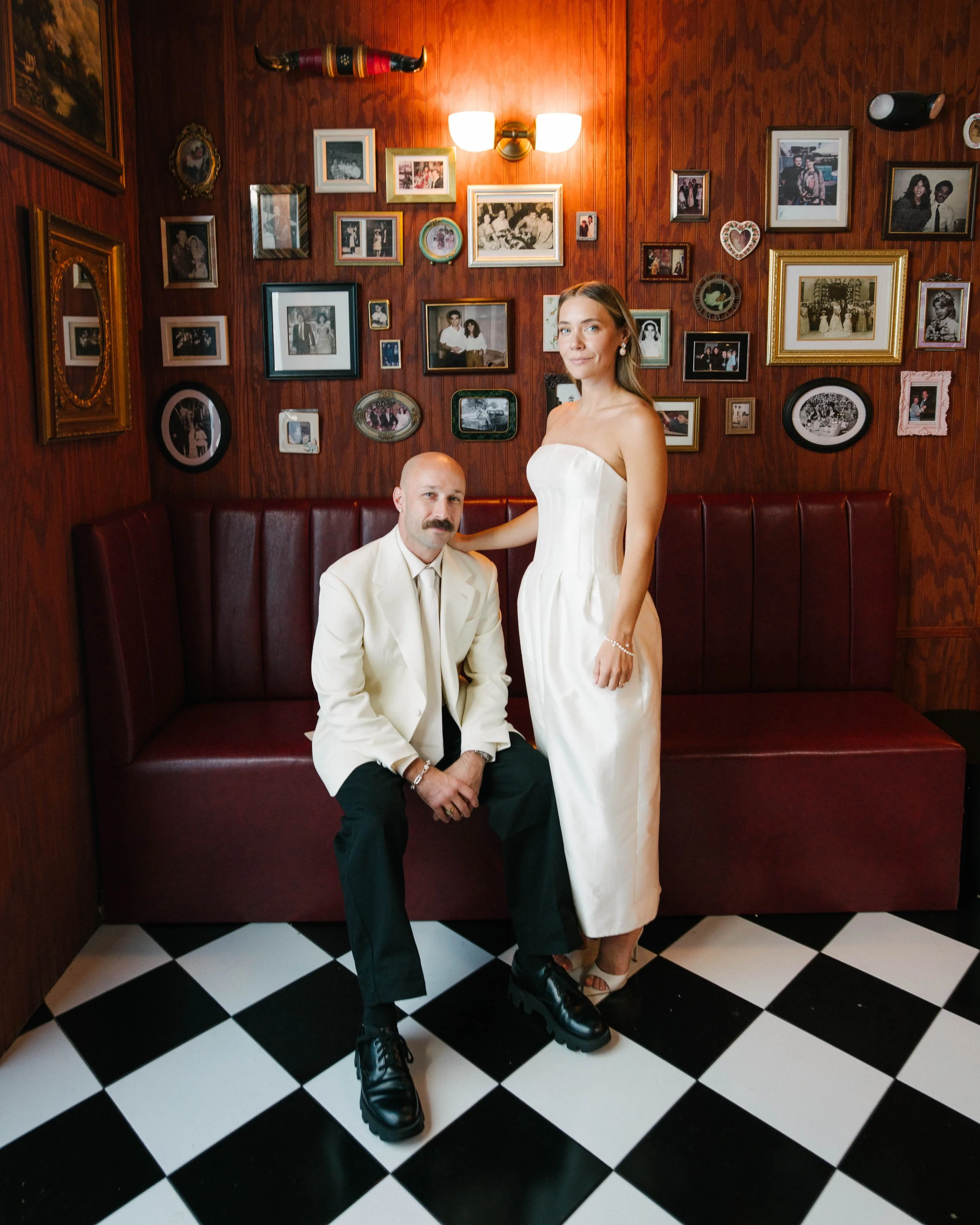 A man and woman posing inside a room with wooden walls, decorated with framed photographs, paintings, and decorative items, including a horn-shaped object. The man is sitting on a red leather bench, wearing a cream suit and black shoes, while the wom