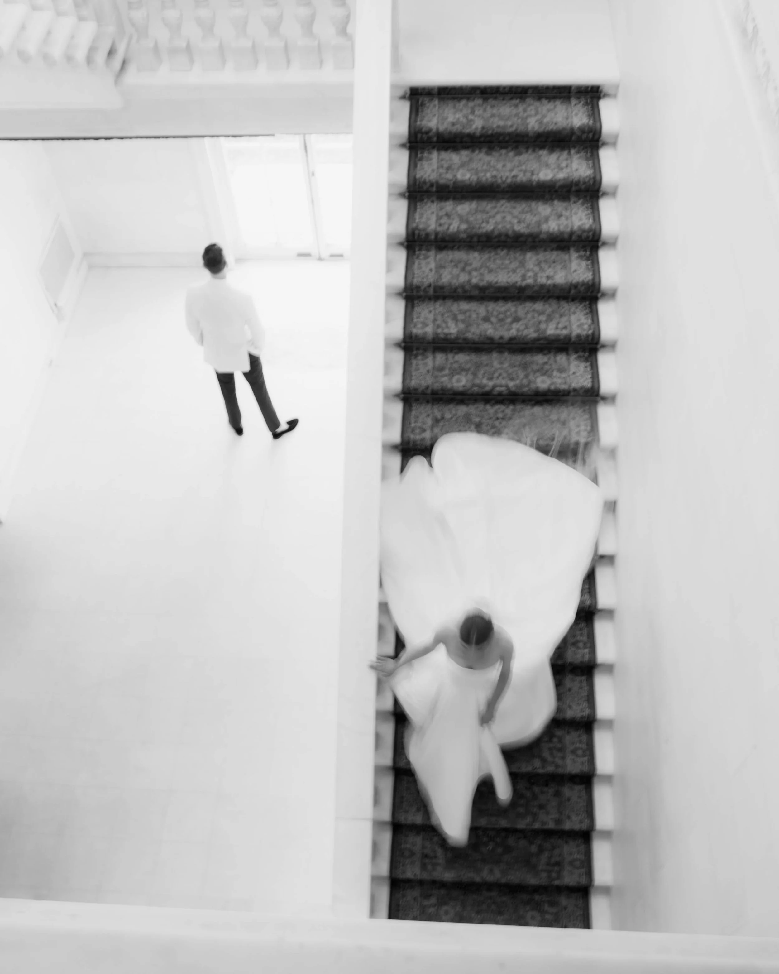 A woman in a wedding dress descending a staircase and a man in formal attire standing at the top of the stairs, viewed from above in black and white.