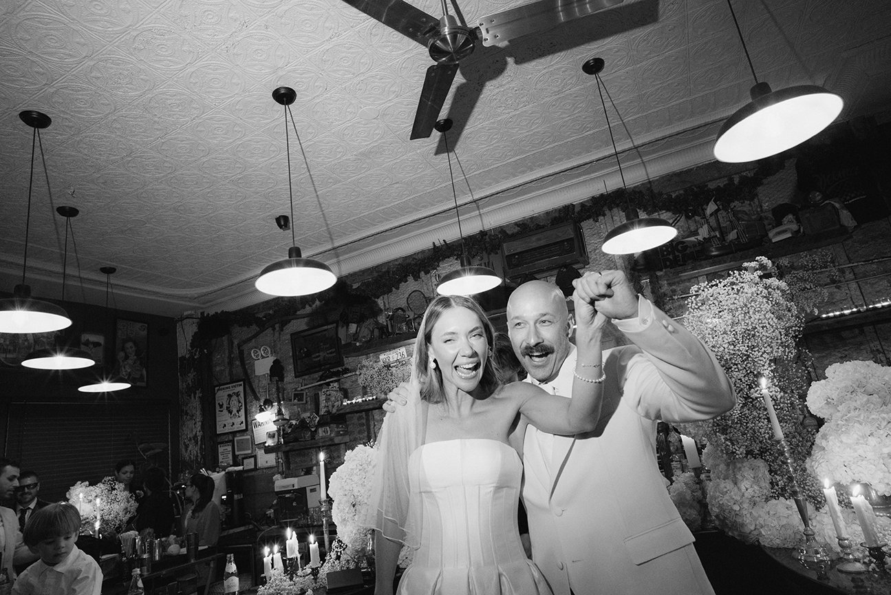 Black and white photo of a bride and groom celebrating at their wedding reception, holding hands and smiling, with flowers and candles in the background.