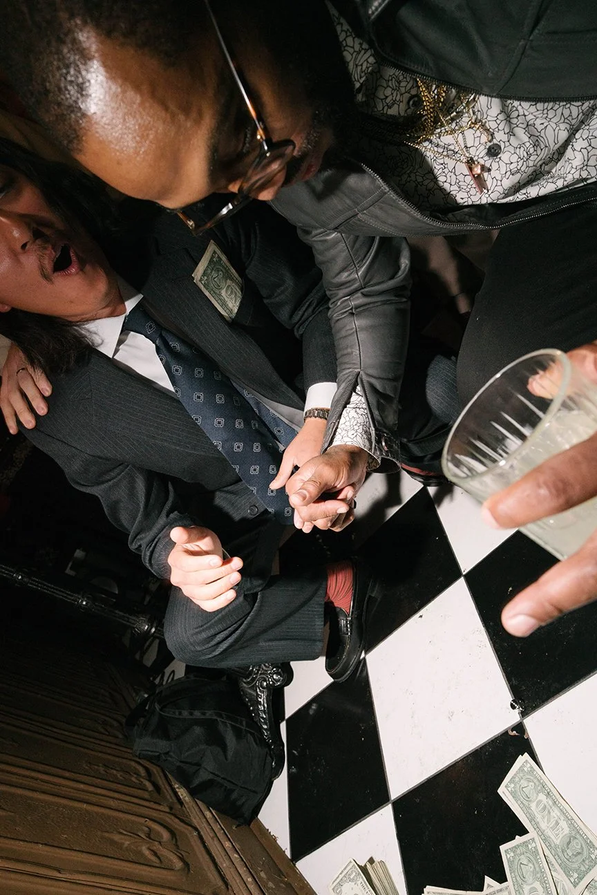 Two men in suits sitting at a table with cash on the floor and a glass of beer, having a conversation or a moment of intimacy, in a setting with black and white checkered flooring.