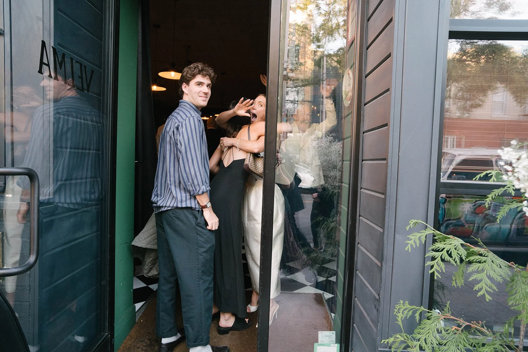 People inside a restaurant seen through a glass door, with one woman making a playful gesture and two men standing nearby.