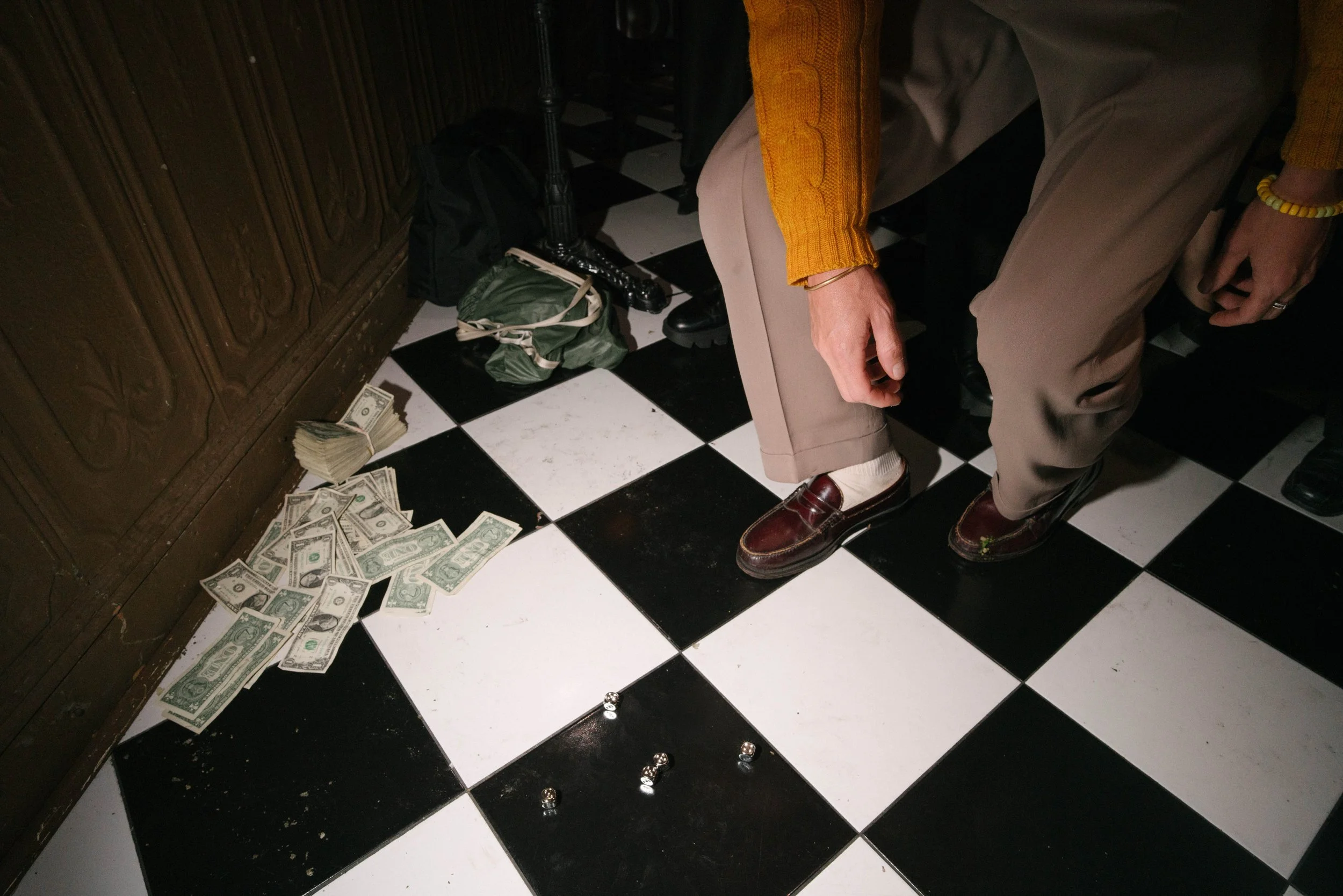 A person wearing a yellow sweater, beige pants, and brown loafers squatting near a pile of cash, some rolled up and some spread on black and white checkered floor tiles. There are small metallic balls or dice on the floor, and a backpack and clothing