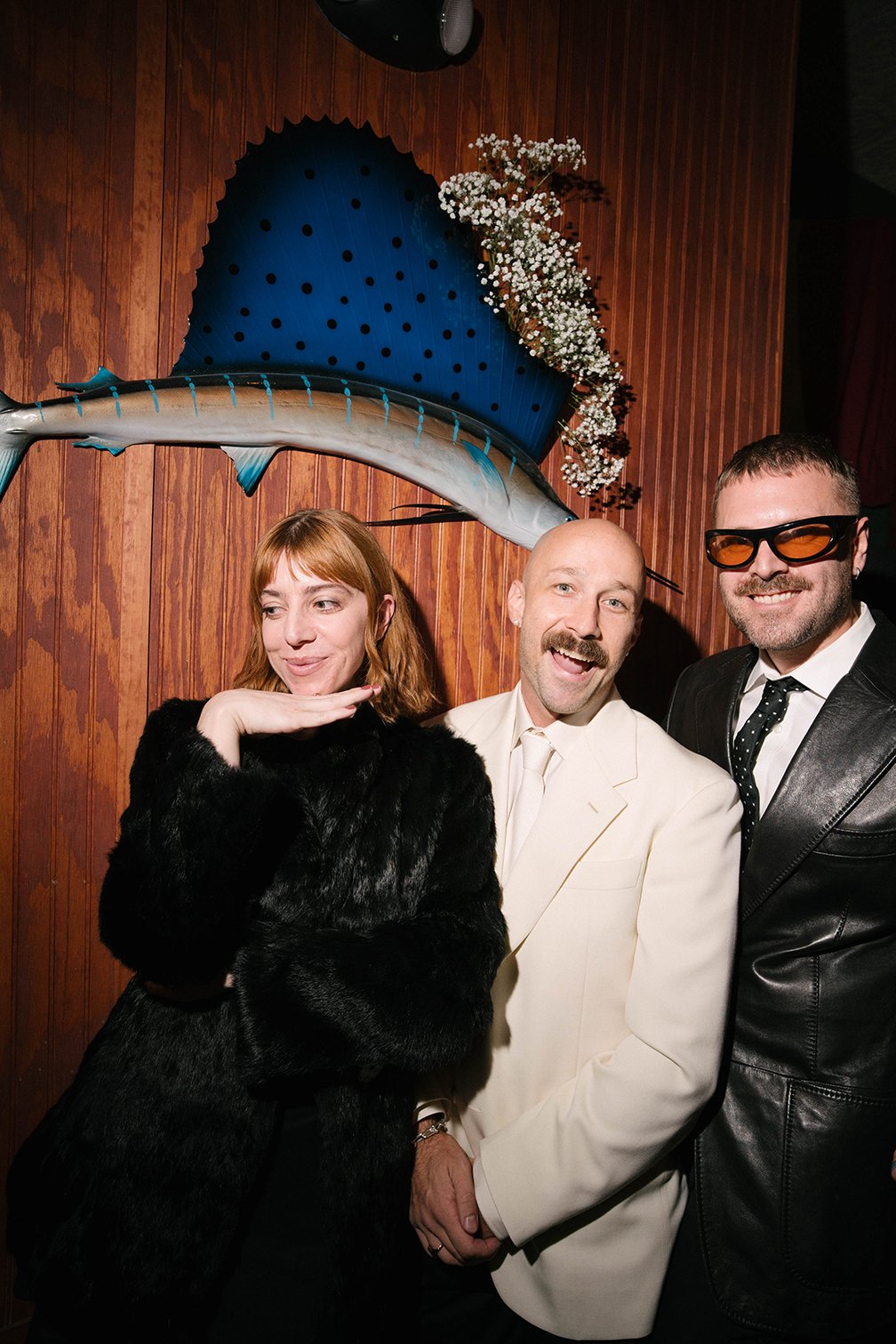 Three people posing in front of a wooden wall with a large fish sculpture and flowers above. The woman on the left has red hair and is wearing a black fur coat, the man in the middle has a bald head, a mustache, and is wearing a white suit. The man o