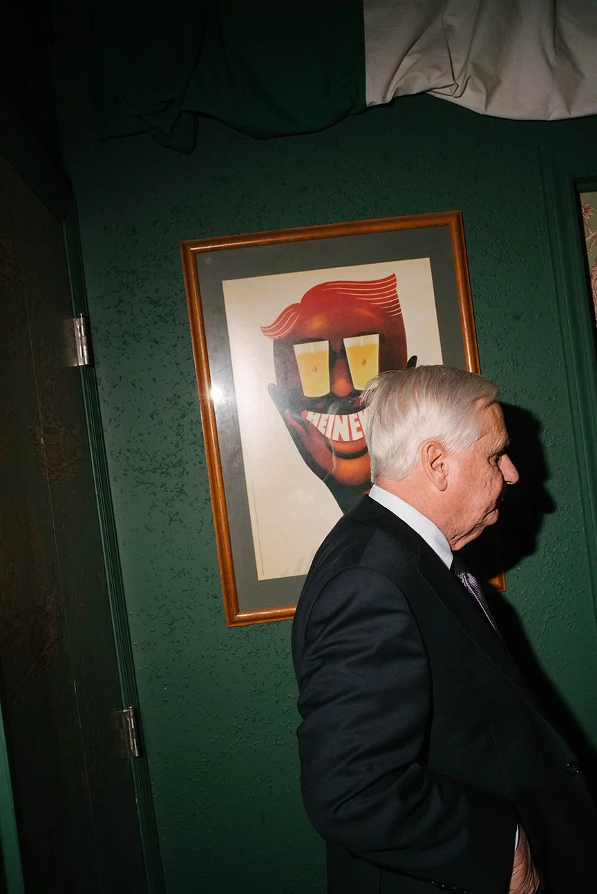An elderly man in a black suit standing in front of a green wall with a framed picture of a cartoon face with glasses, a mustache, and a painted smile.