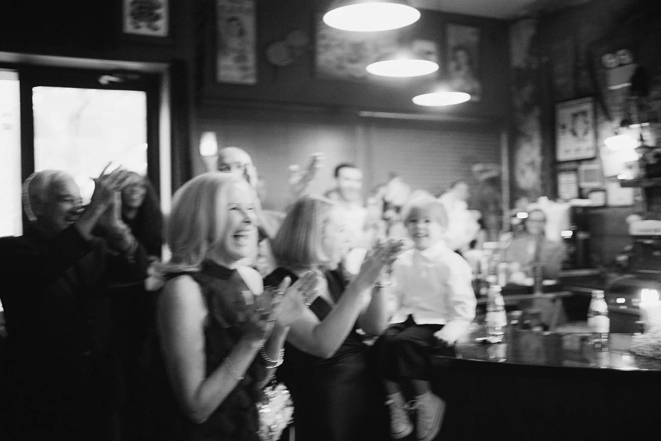 Group of people celebrating and smiling in a restaurant or bar, clapping and enjoying a festive moment.