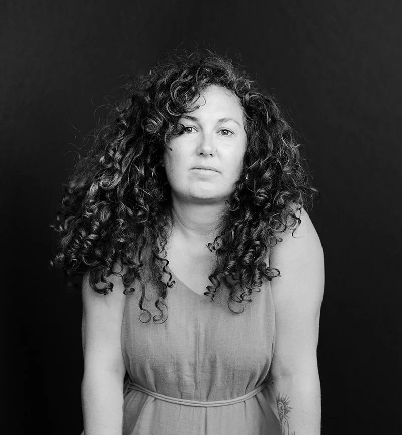 Black and white portrait of a woman with curly hair wearing a sleeveless dress against a dark background.