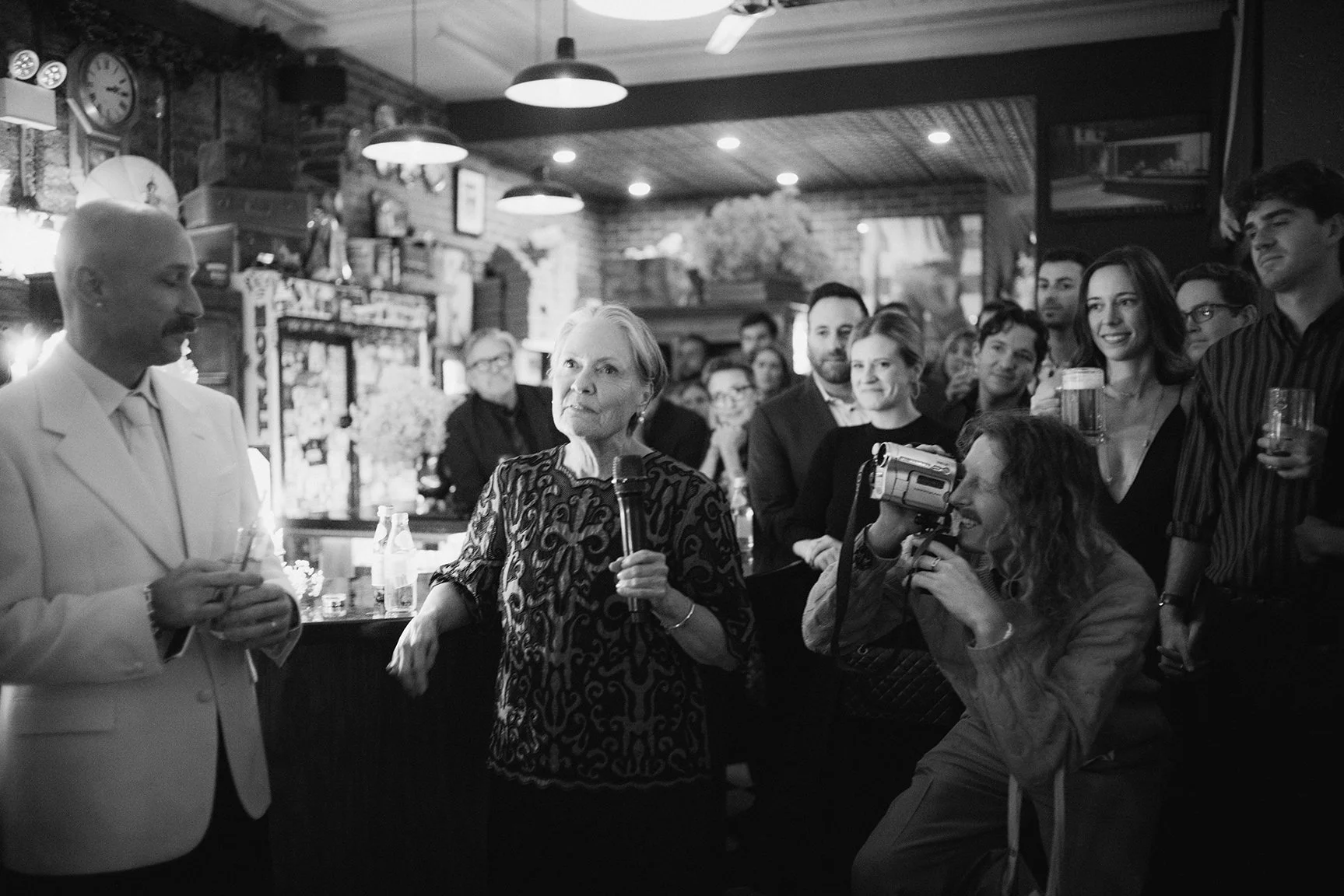 A woman holding a microphone addressing a group of people in a bar, with a woman filming her and others smiling and listening.