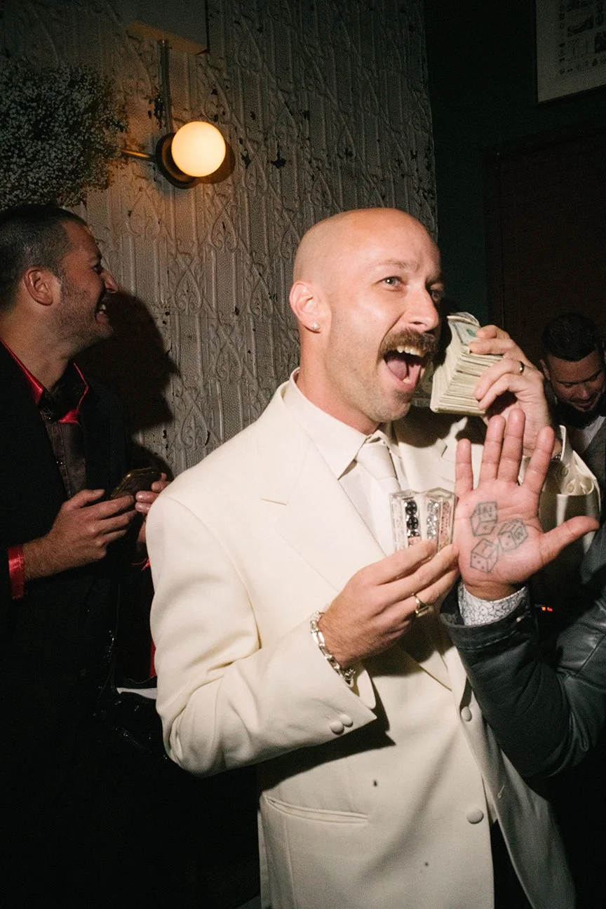A man with a bald head, mustache, and earrings, dressed in a light-colored suit, holding a stack of cash to his ear as if talking on the phone, smiling and raising his other hand with a tattoo of dice, in a dimly lit indoor setting with other people 