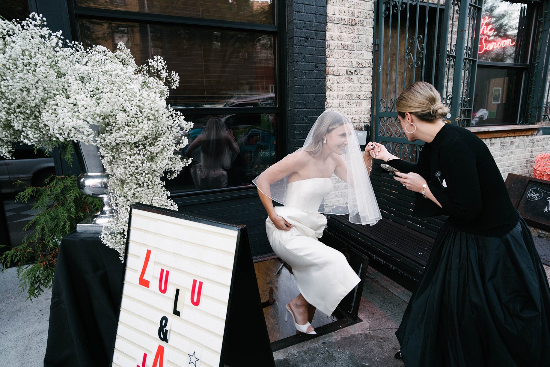 A woman in a white wedding dress and veil sitting on a bench outside a building, smiling as another woman in black dress and large hoop earrings holds her hand and uses a phone. There are flowers and a signboard nearby.
