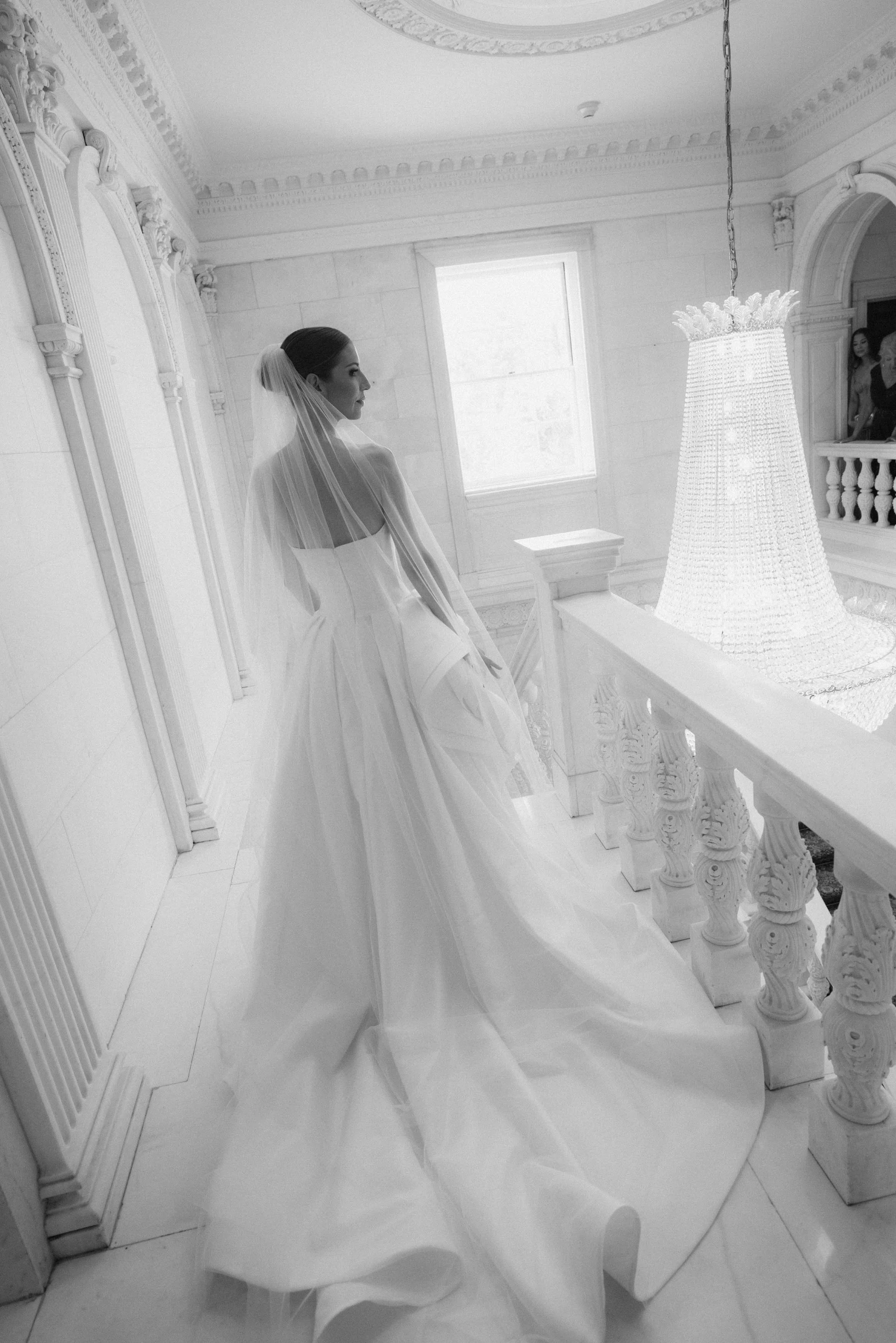A bride in a wedding gown and veil standing on a marble staircase in a bright, ornate interior with intricate moldings and a large chandelier.