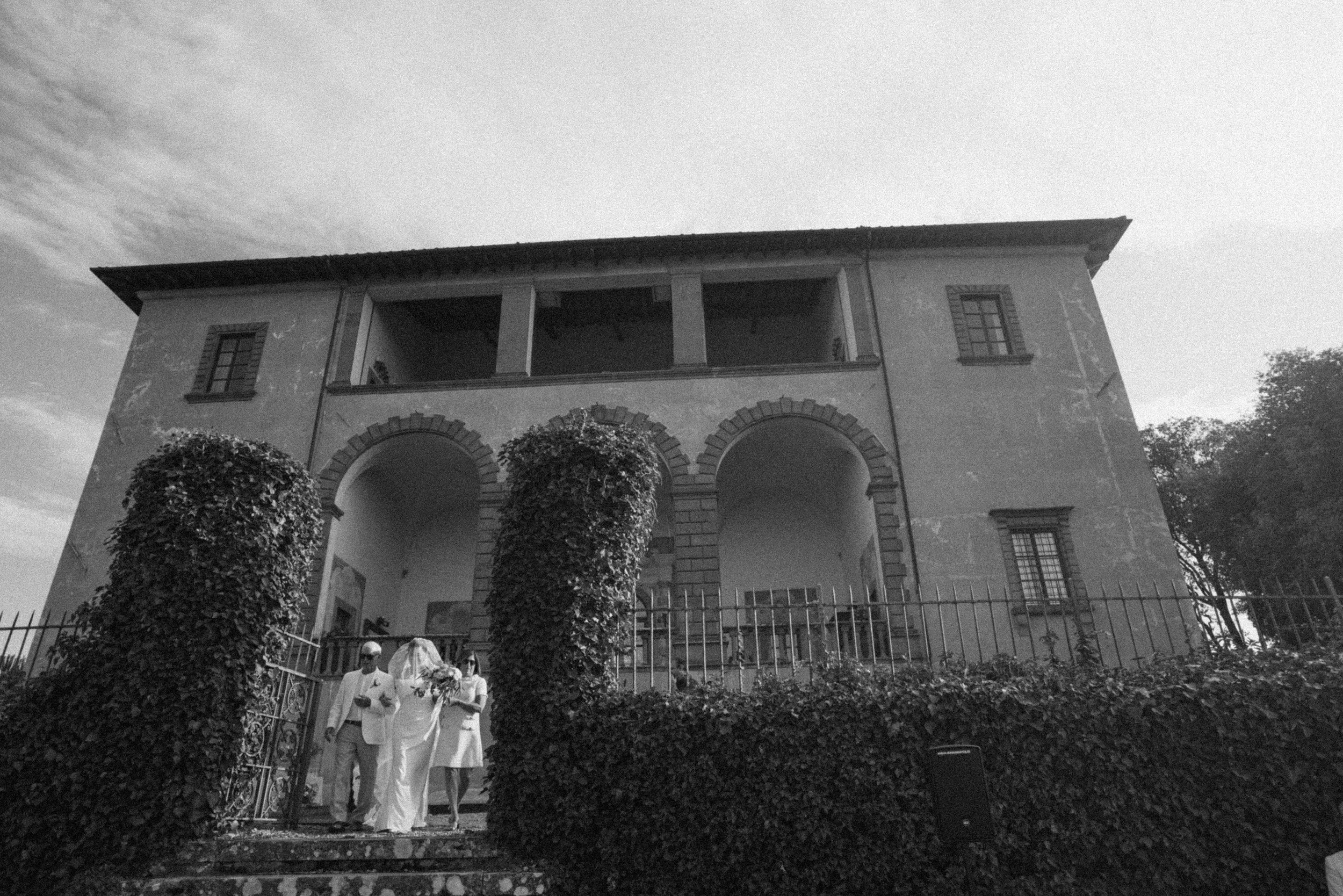 A bride whos face is covered by a veil is walked down the aisle by her parents walk in front of a historic villa in Tuscany Italy