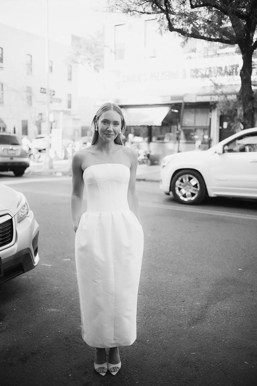 A woman in a strapless white dress and high heels stands on a city street with cars and buildings in the background.