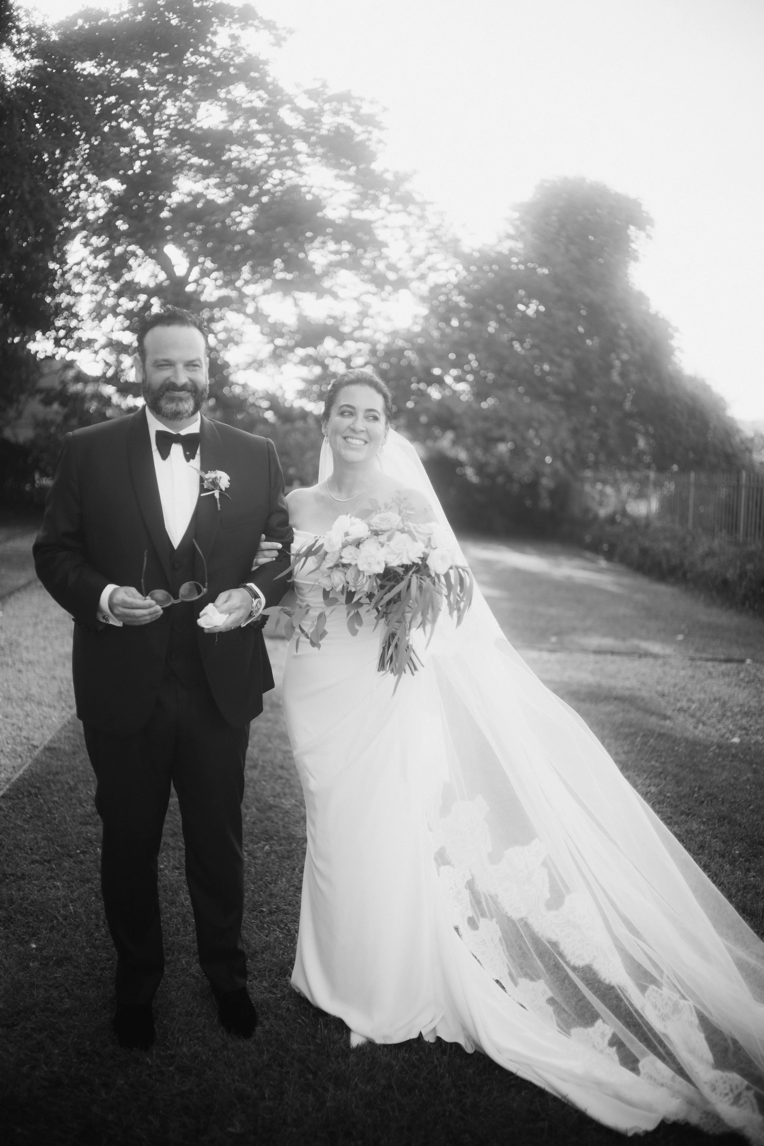 Black and white photograph of a bride and groom walking outdoors, smiling, with trees in the background. The bride is dressed in a wedding gown with a veil and holding a bouquet, while the groom is wearing a tuxedo with a bow tie and holding sunglasses.