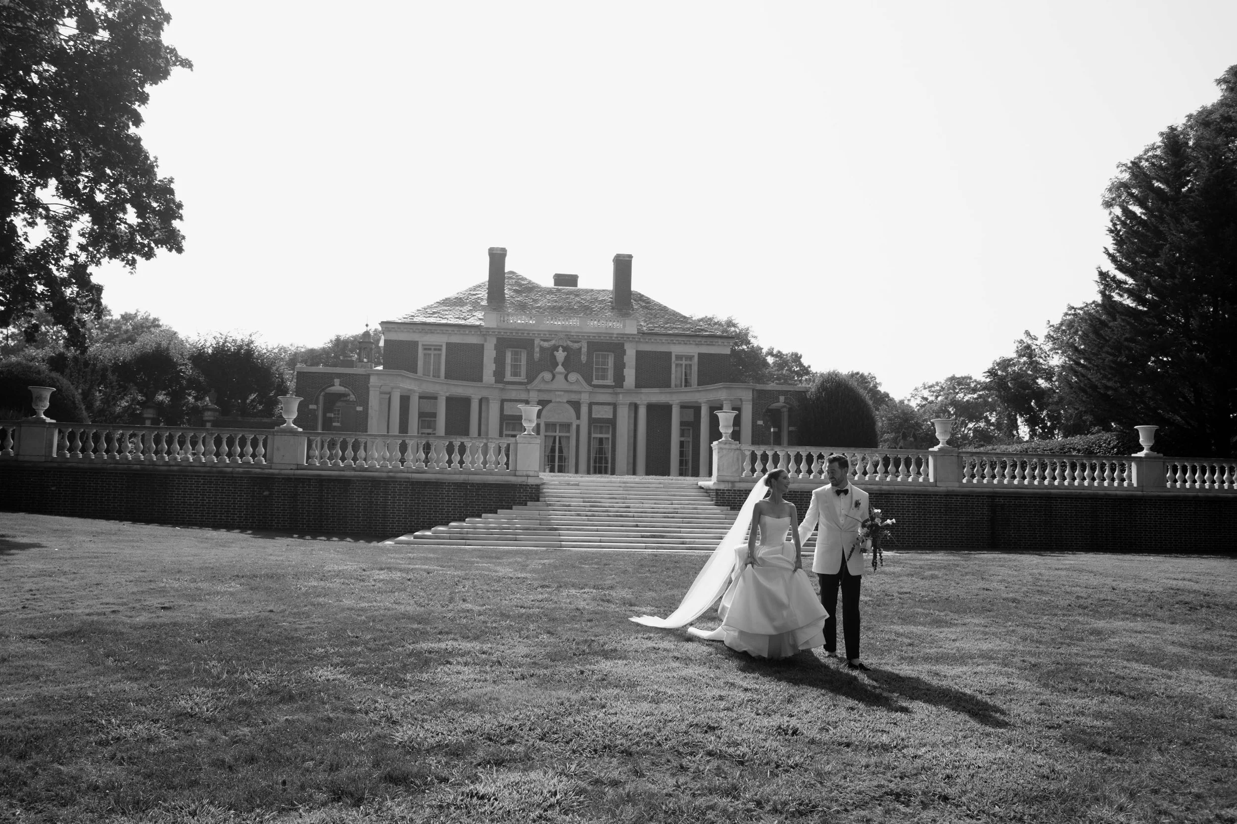 A bride and groom walking on a lawn in front of a large historic mansion, with the bride wearing a wedding dress and veil, and the groom in a tuxedo, holding a bouquet.