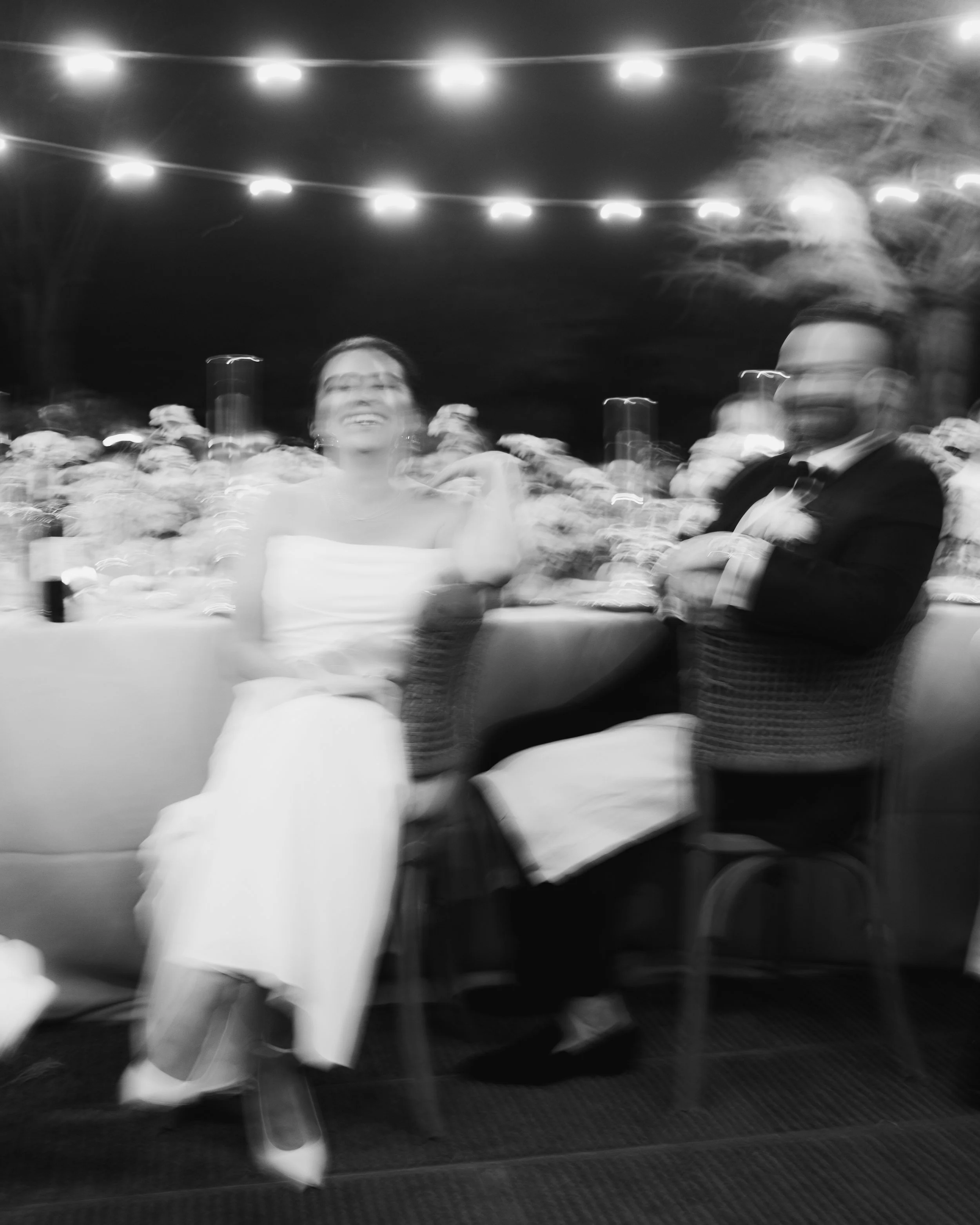 A black and white photo of a bride in a strapless dress and a groom in a tuxedo sitting at a table during a wedding event at night. The background has string lights and a crowd of people. Villa Mangiacane in Tuscany Italy.