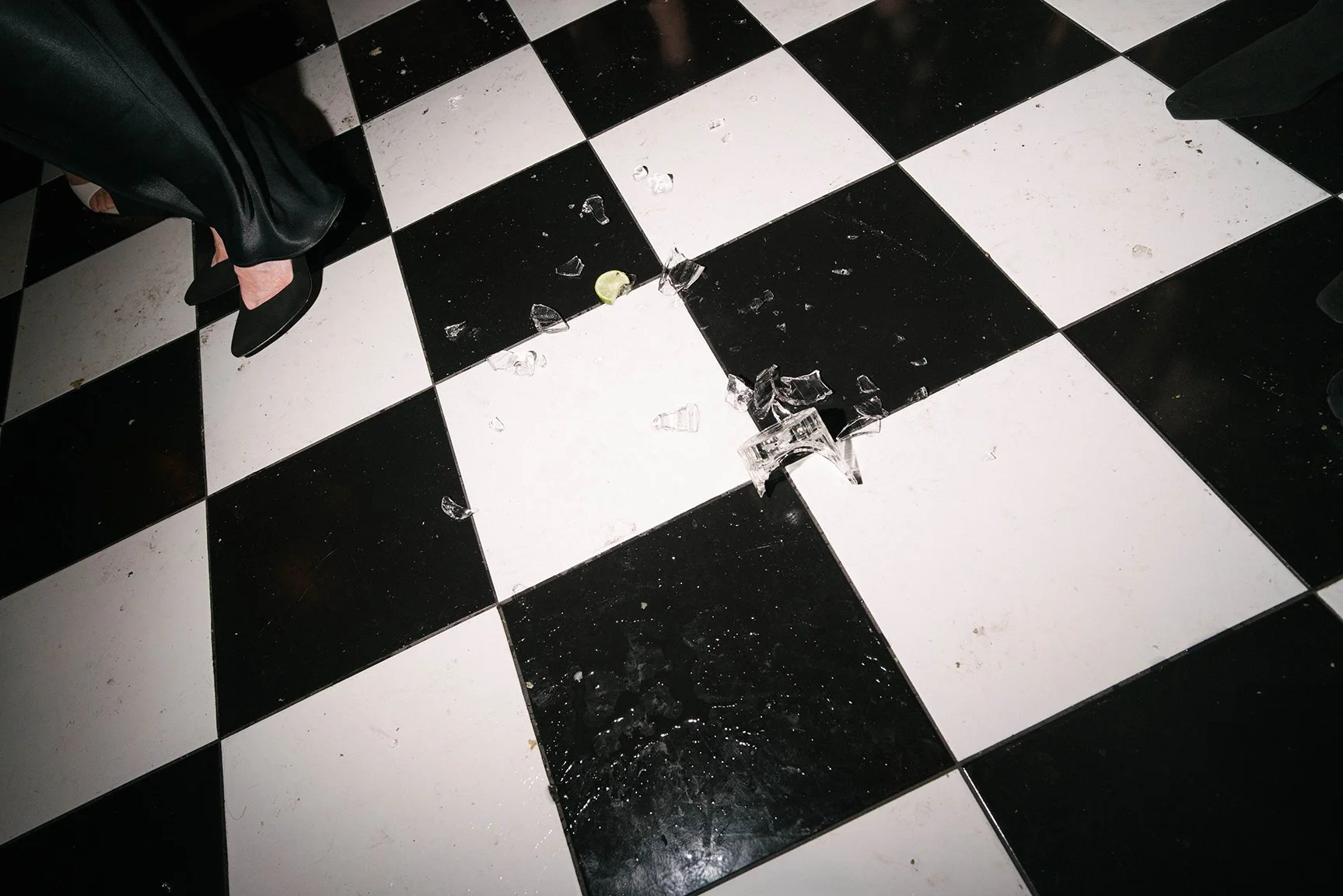 Broken glass on a black and white checkered floor, with a woman in black trousers and heels standing nearby.