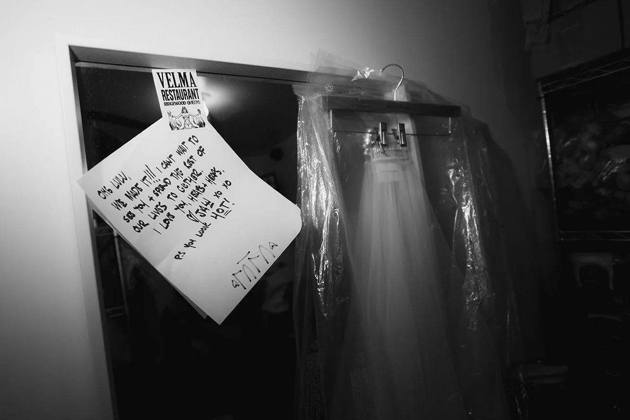 Love note posted on a mirror, expressing love for someone and saying they can't wait to see them again. The note is from someone named Ellie. A wedding dress hangs on a hanger covered with plastic in the background.
