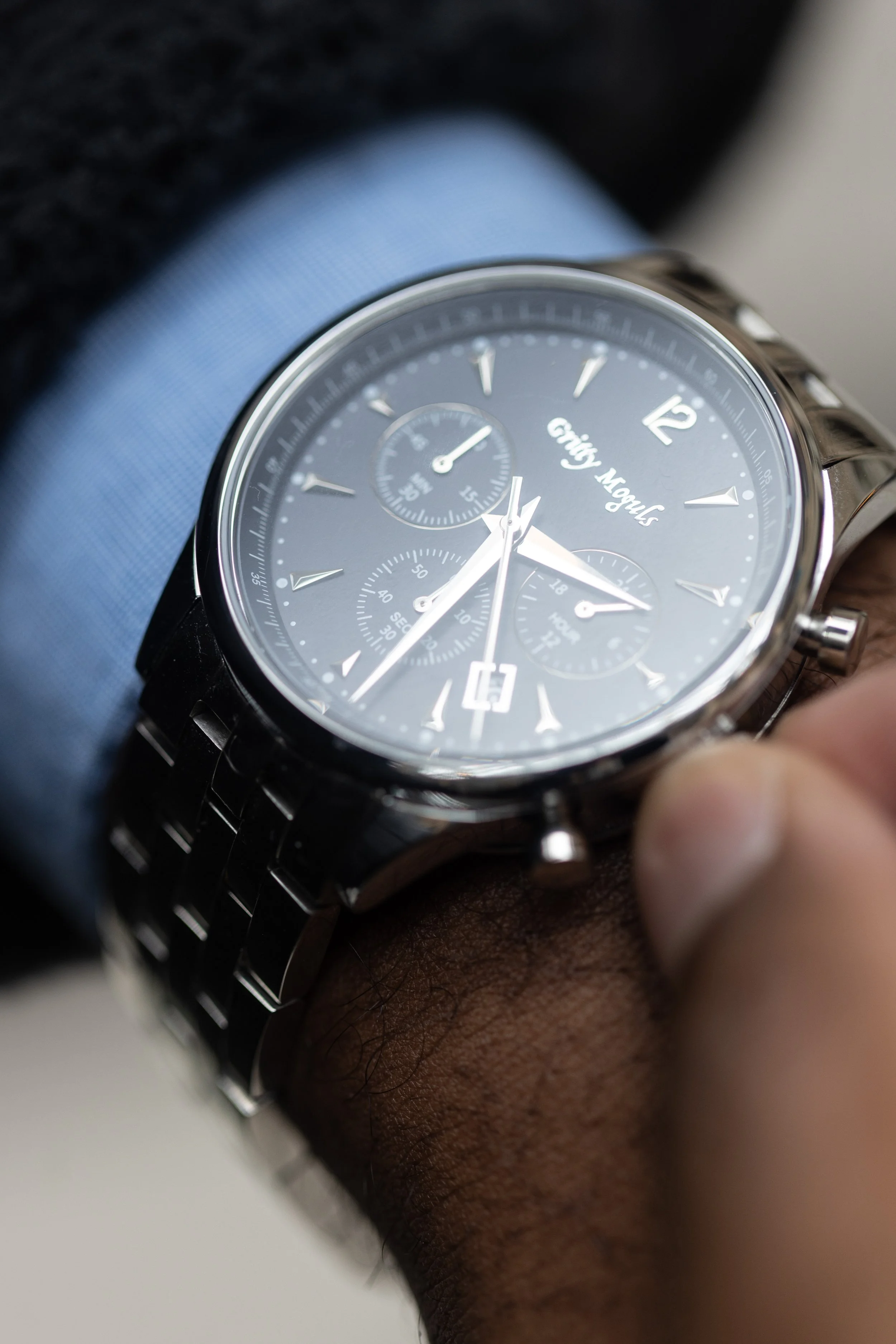 A close-up of a man's wrist wearing a black metal watch with a dark face and three subdials, showing the brand name 'Fifty Moul'.