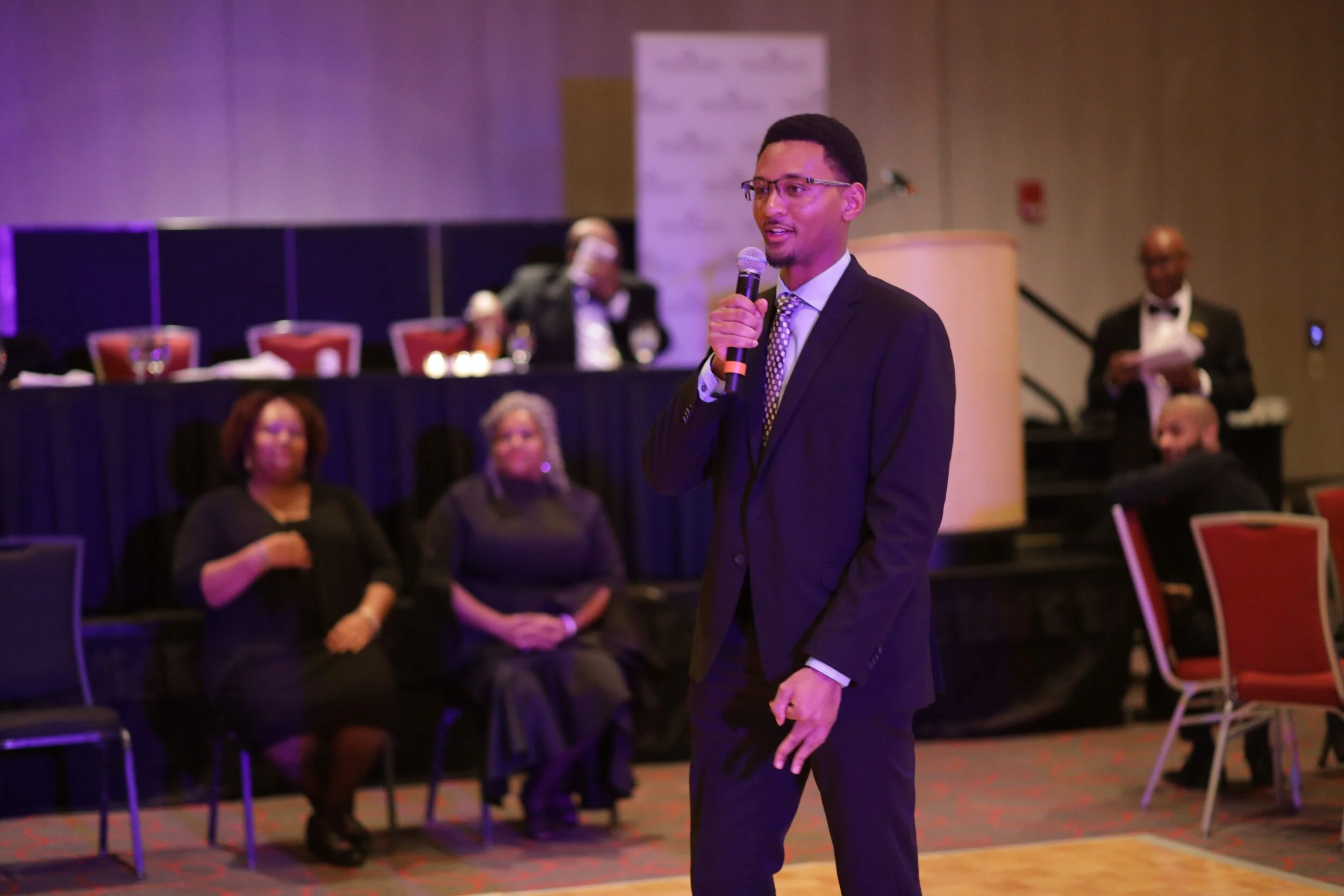 A man in a black suit and glasses speaking into a microphone at a formal event, with an audience seated in the background.