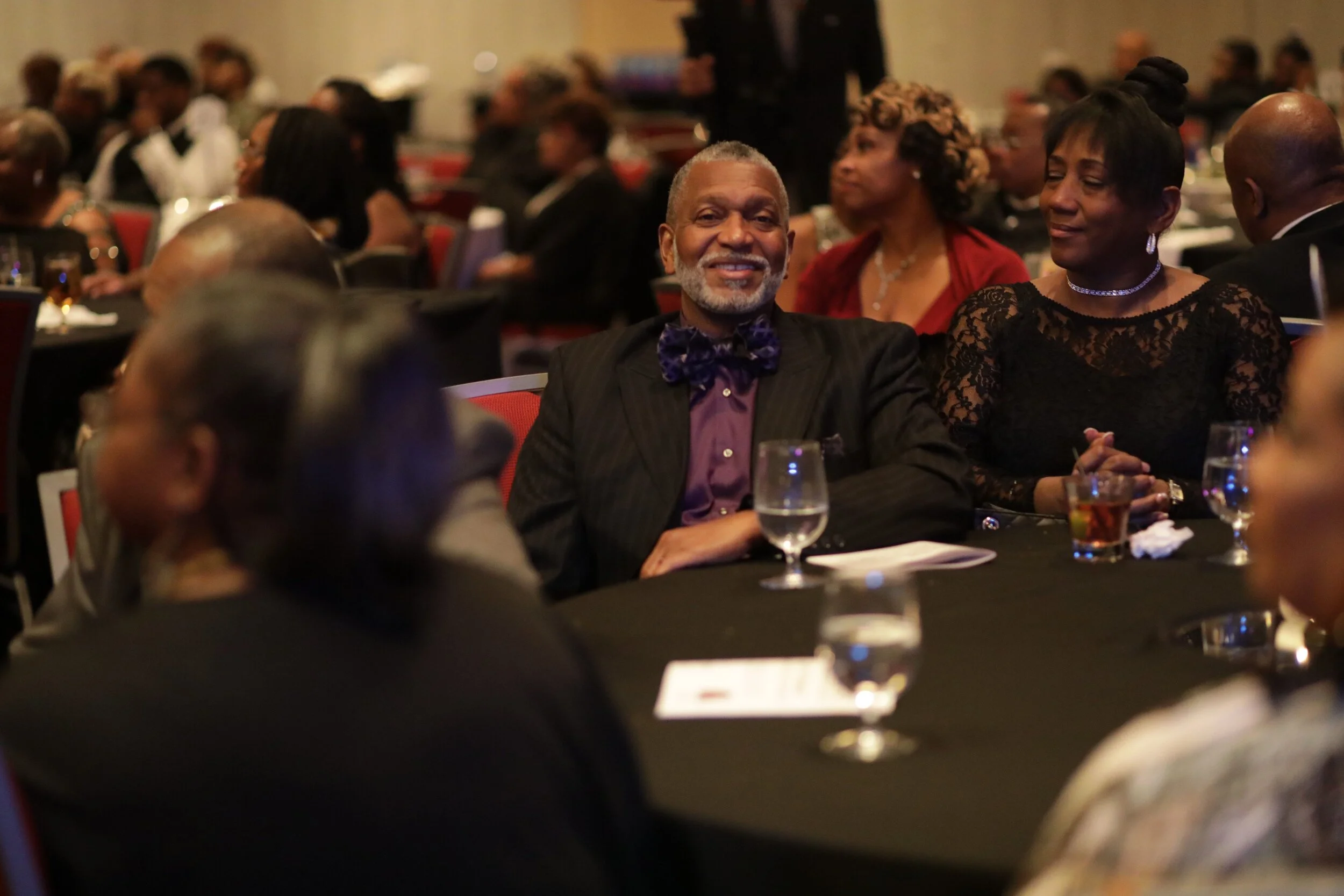 A smiling man in a suit with a purple shirt and bow tie sitting at a banquet table with others in formal attire.