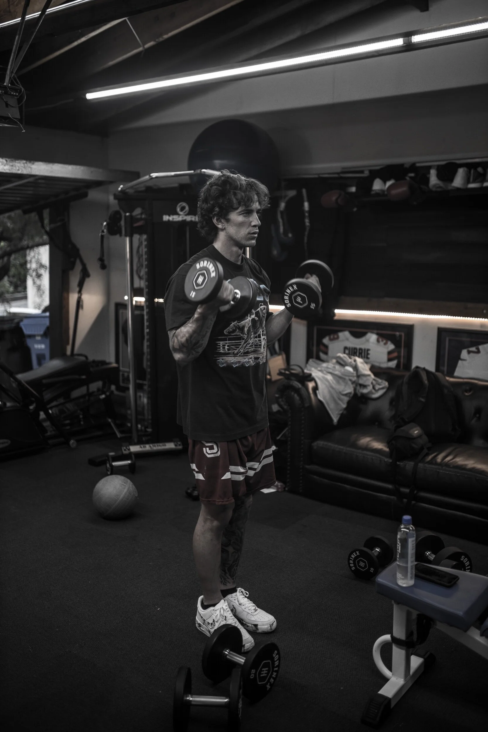 A man lifting dumbbells in a gym, black and white photo, with various workout equipment and framed sports jerseys on the wall.