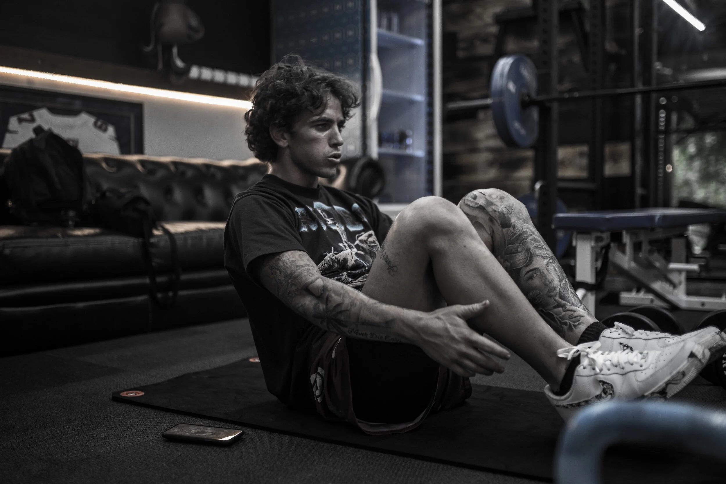 A young man with tattoos on his arms and legs doing sit-ups in a gym, sitting on a mat with a phone nearby.
