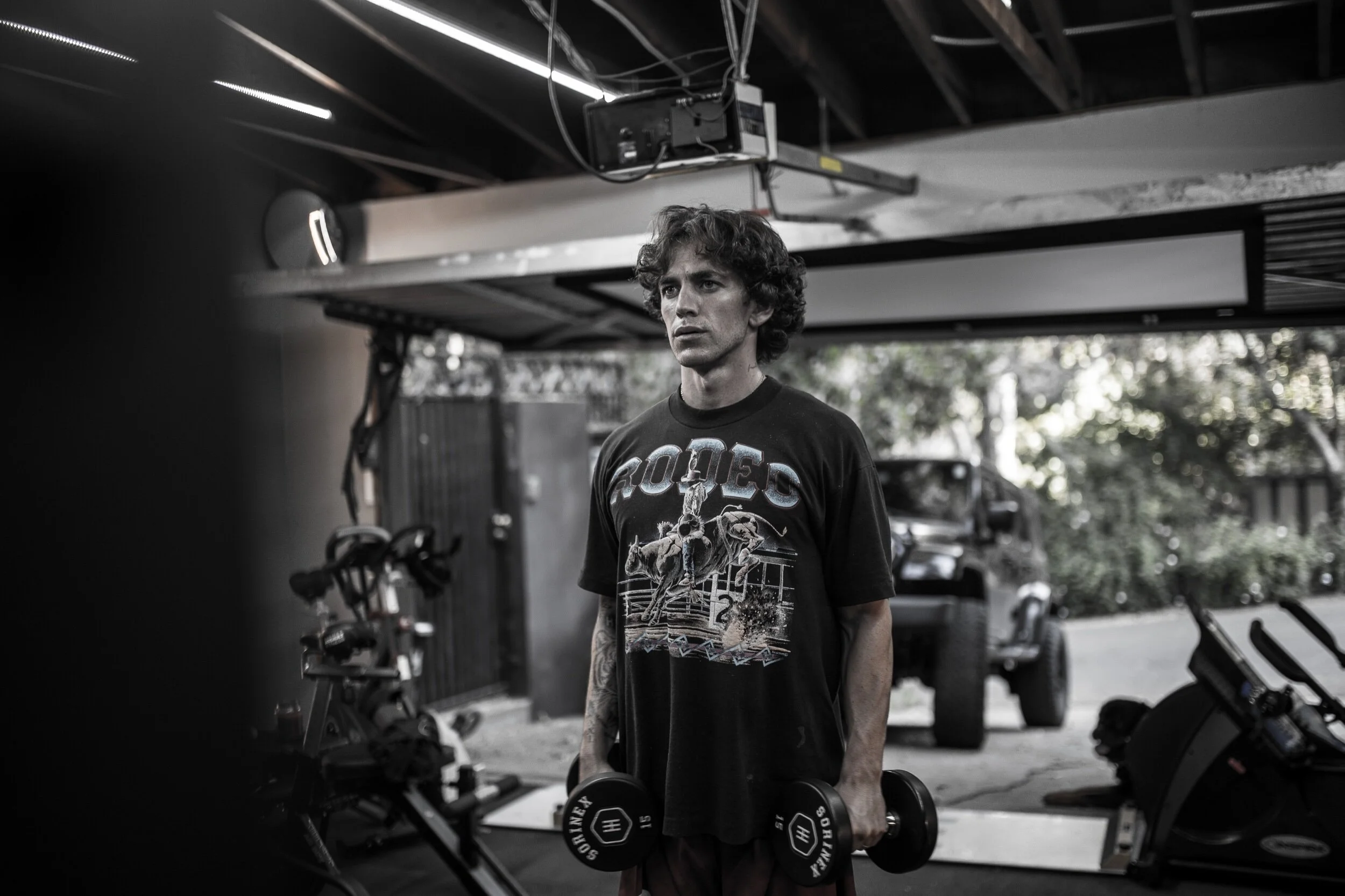 A man holding dumbbells in a garage gym with exercise equipment and a vehicle in the background.