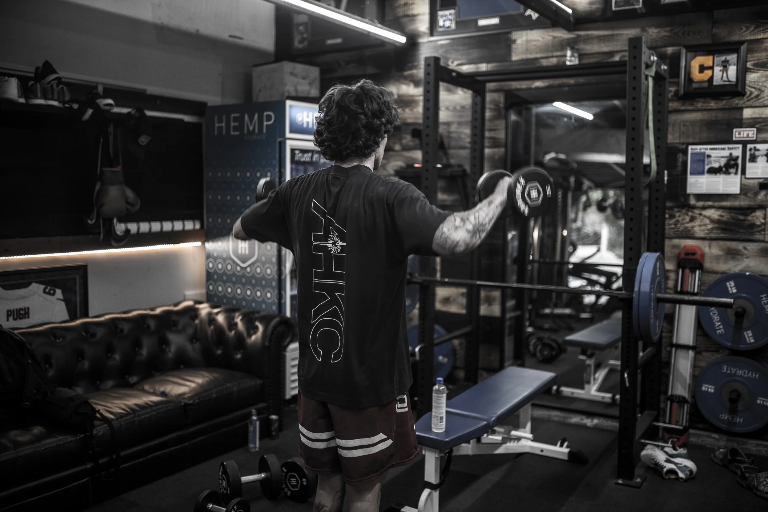 A person with curly hair and tattoos on their arm is lifting a dumbbell in a gym, facing away from the camera, with workout equipment, weights, and a black leather couch in the background.