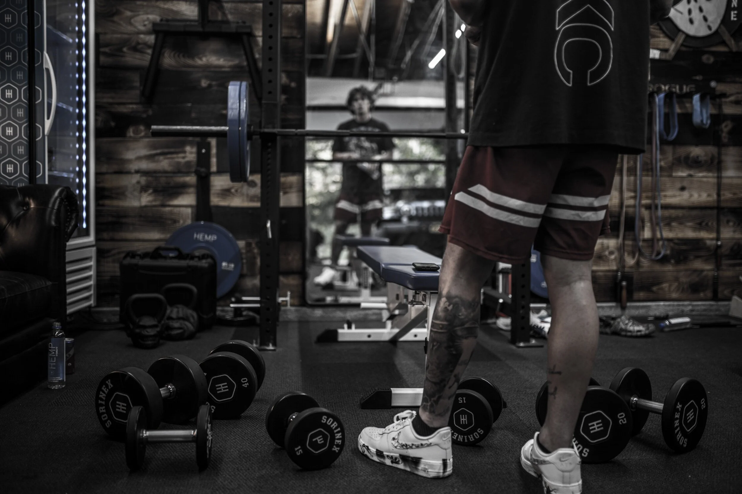 A person standing in a gym with various weights on the floor, including dumbbells and kettlebells, with another person visible in the background through a mirror.