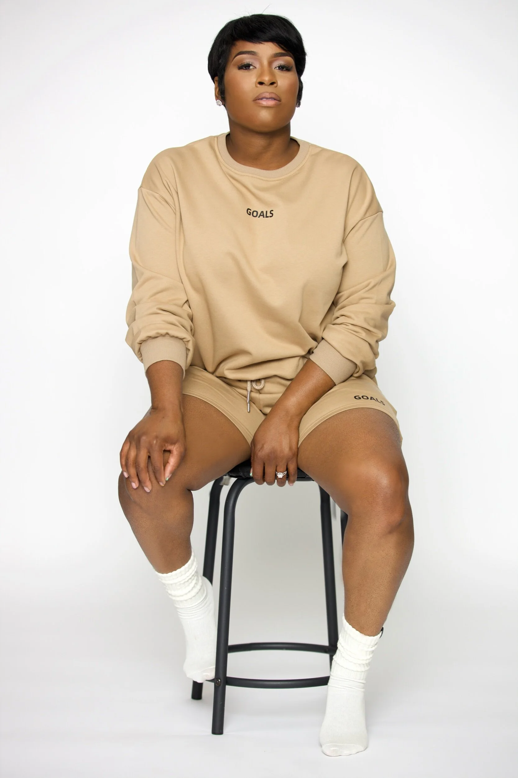 A woman sitting on a black stool, wearing a beige sweatshirt and shorts with the word 'GOALS' printed on them, and white socks, against a plain white background.