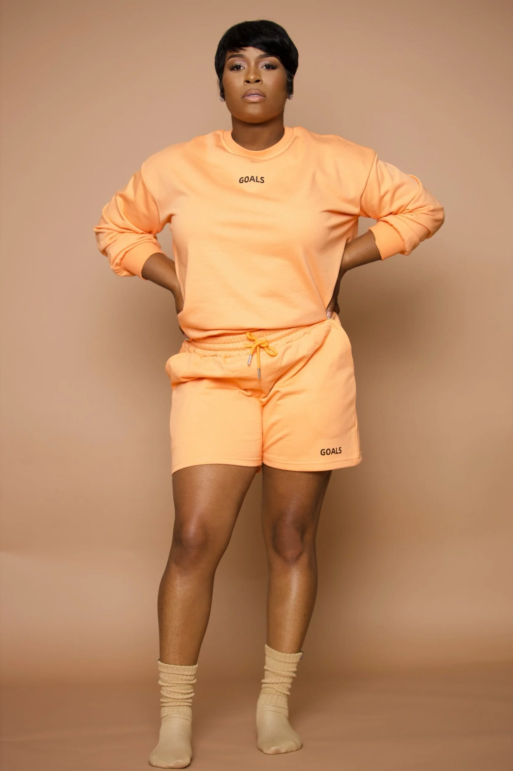 A woman with short black hair wearing a peach-colored sweatshirt and matching shorts, standing against a beige background.