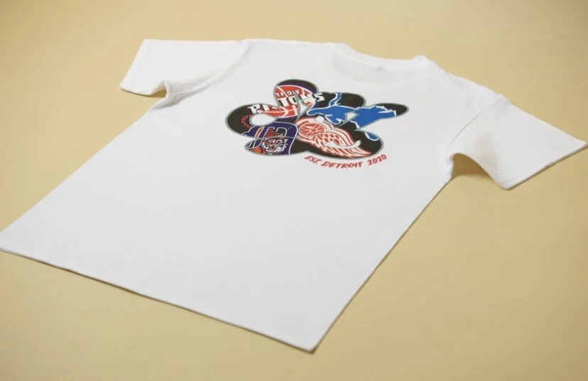 White T-shirt with a cluster of sports team logos and a red text that says 'Detroit 2020' lost on a beige surface.