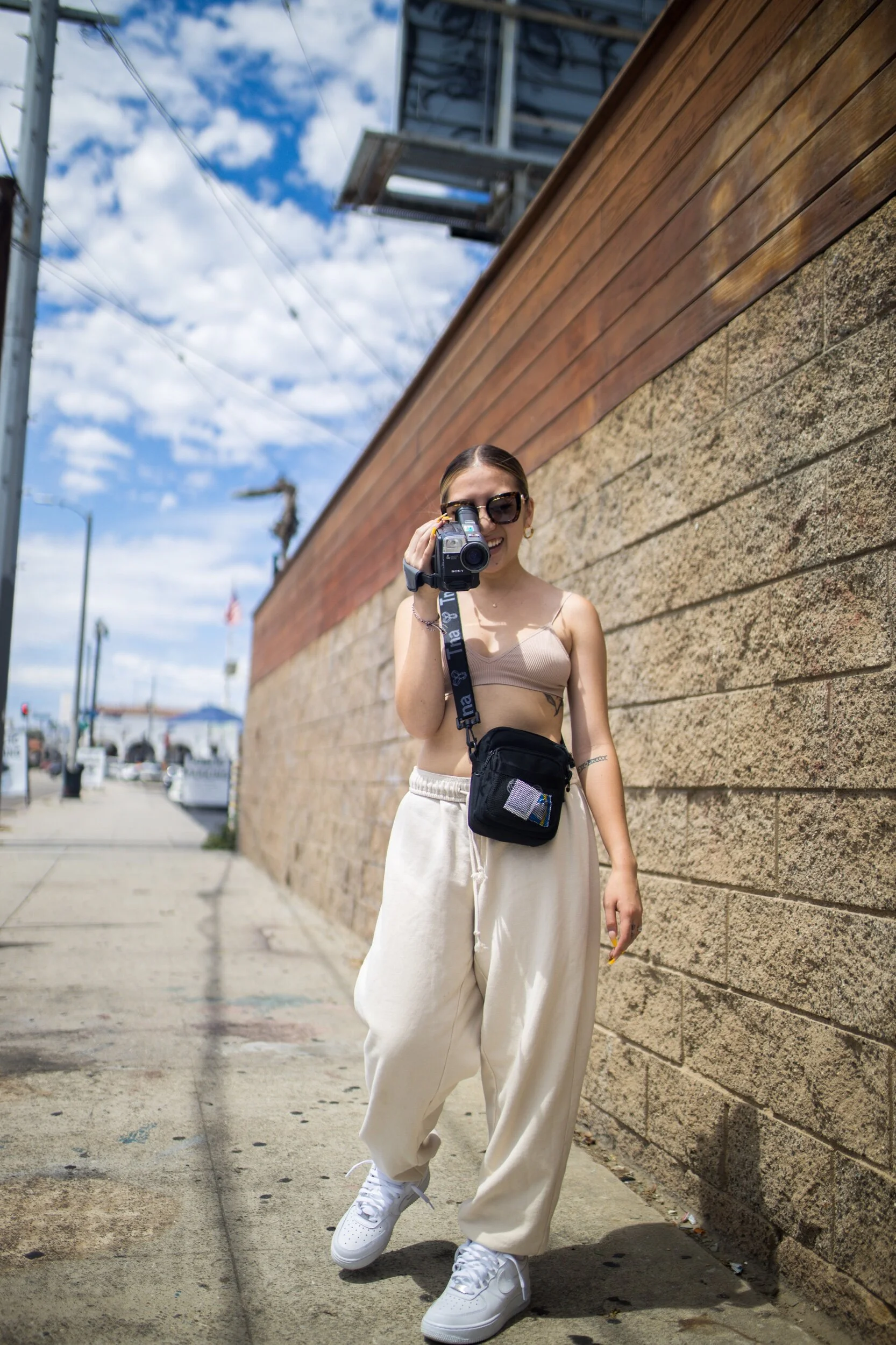 A woman with sunglasses and a beige crop top standing outdoors on a sidewalk, holding a camera pointed towards the viewer, with a brick wall behind her and a partly cloudy blue sky.